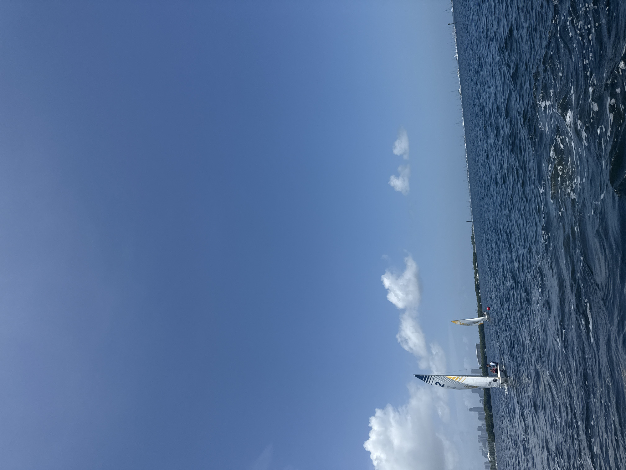The image depicts a bright, clear day with a vast expanse of deep blue sky and dark blue, choppy water. The scene is oriented vertically, with the horizon running from the bottom left to the top right. Several sailboats are visible on the water, engaged in what appears to be a race.
In the foreground, a sailboat with a white hull and sails marked with blue and yellow stripes is prominent. The number "2" is clearly visible on its sail. Two people are on board this sailboat, seemingly steering and managing the sails. Further in the distance, another sailboat, primarily white with yellow accents, can be seen. A small red flag is visible on this boat.
The background reveals a distant cityscape, suggesting the activity is taking place in an urban coastal area. The buildings form a faint, hazy line along the horizon. Scattered white clouds drift across the sky, some appearing fluffy and others more wispy. The sunlight is strong, casting sharp reflections on the water's surface and highlighting the texture of the waves. The overall impression is one of a lively daytime sailing event under fair weather conditions, likely in a location such as Miami, given the city skyline. No discernible emotions are conveyed, and the focus is on the activity and the natural elements.