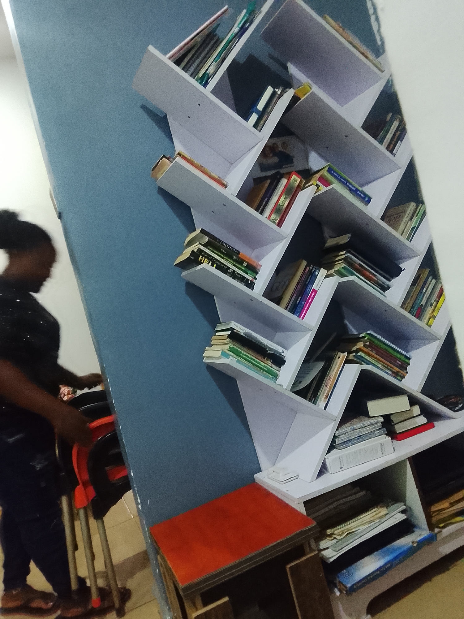 The image depicts an indoor scene in Karmajiji, Nigeria, dominated by a large, white, zig-zag-patterned bookshelf mounted on a blue wall. To the left, a dark-skinned female individual is partially visible, out of focus.
The individual, positioned on the left side of the frame, is seen from the mid-torso down. She has dark hair styled in a bun or tied back and is wearing a dark, possibly sequined or textured, long-sleeved top and dark blue distressed jeans. Her hands are positioned near a stack of two chairs with black seats and backrests and orange-red metal frames. Her feet, clad in brown flat shoes or sandals, are visible on a light-colored tiled floor.
The prominent white bookshelf occupies the right two-thirds of the image. It features multiple shelves arranged in an angular, interconnected "Z" or chevron pattern, appearing to be built into the blue wall behind it. The shelves are filled with numerous books of varying sizes, colors, and thicknesses, stacked both horizontally and vertically. Visible book titles on the shelves include "EMOTIONS," "Role of Engagement," and "HELL" on the upper left section. On a lower shelf, a book with the word "BITCHES" is discernible. Below the central section of the bookshelf, a small table or stool with an orange-red top and dark frame is positioned on the tiled floor. Directly beneath the lowest shelves, a blue folder or binder is visible among other stacked items.
The blue wall behind the bookshelf extends across the frame. To the far left, a light-colored wall or doorway is visible, indicating an interior corner. The floor is covered with light-colored tiles. The scene is illuminated by what appears to be ambient artificial light, contributing to a slightly warm, yellowish cast. The overall image has a slight tilt, making the vertical elements appear to lean clockwise.