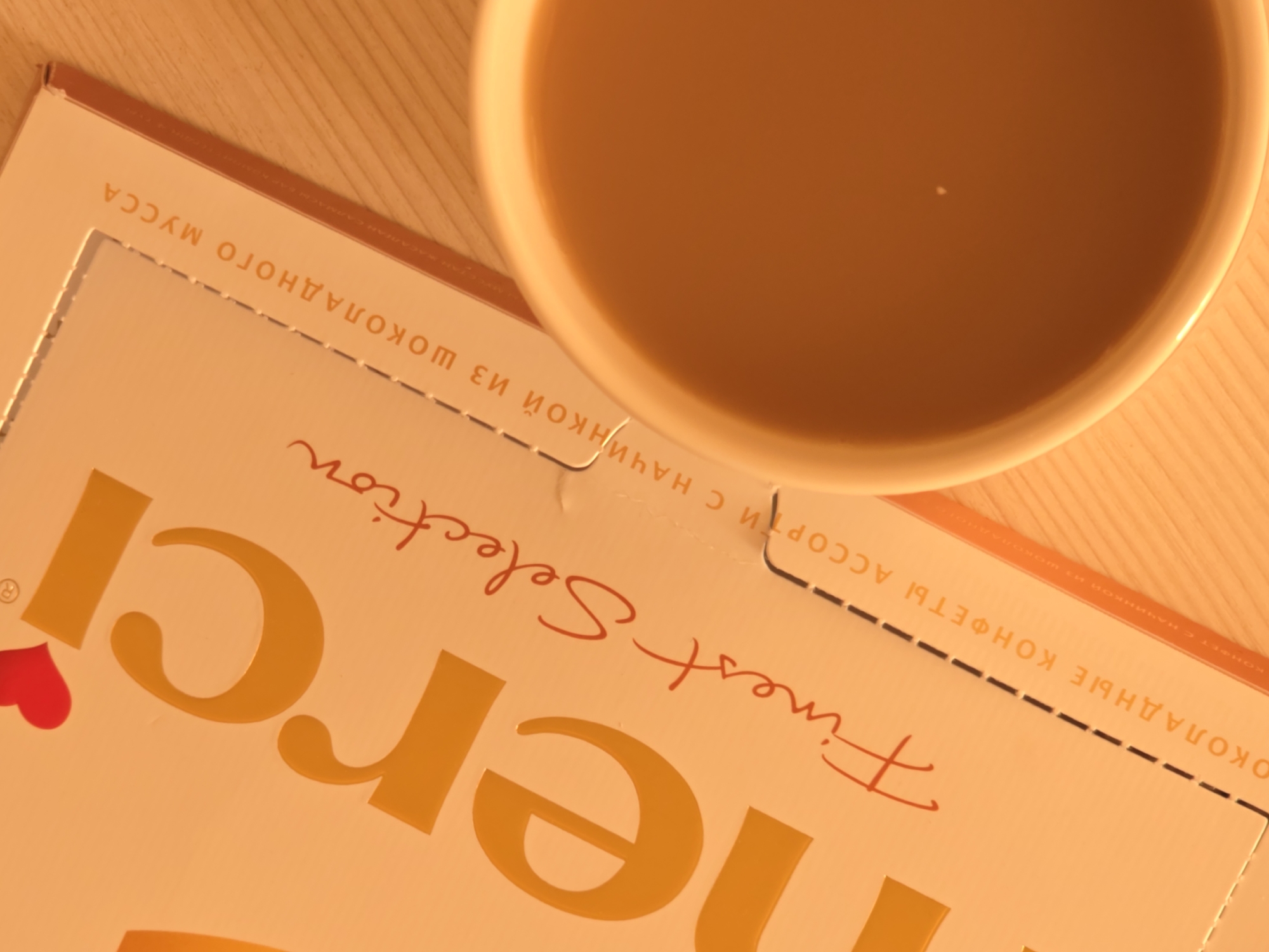 The image shows a close-up overhead view of a white porcelain mug filled with a light brown beverage, possibly coffee or tea, placed on a wooden surface. A small, bright white speck, possibly a reflection or a tiny air bubble, is visible in the center of the mug's surface. To the left of the mug is an open orange cardboard box of Merci chocolates. The box has bold, yellow embossed lettering that reads "merci" and below that, in red script, "Finest Selection." Further down the box, in smaller yellow lettering, are Russian words that translate to "chocolate candies assortment with chocolate mousse." The lighting casts a warm glow over the scene, suggesting indoor lighting. The wooden surface appears to be a table with a visible wood grain pattern. The image is cropped such that only a portion of the mug and the box are visible. There are no people or animals in the frame. The scene depicts a casual, quiet moment, possibly a breakfast or a break. The location of Rostov-on-Don, Russia is not visually ascertainable from the image itself.