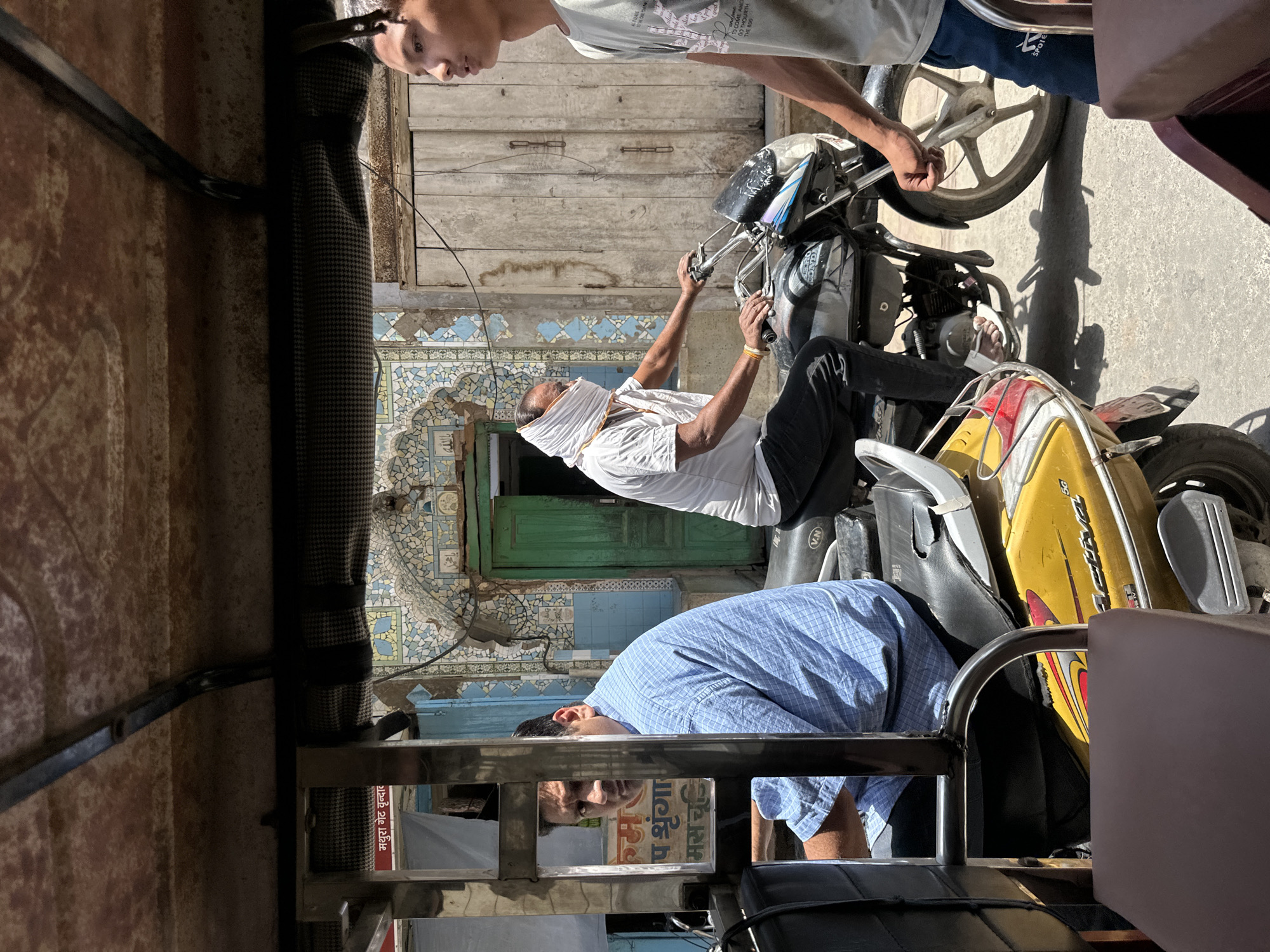 Captured from inside a vehicle, the image depicts a street scene in Vrindavan, India. On the street, a person wearing a white shirt and a head covering works on the handlebars of a motorcycle, while another person watches. Above them, a young man is touching the wheel of the motorcycle. In the background are buildings with decorative blue and white tiled exteriors and green doors. The view from inside the vehicle, includes two seated passengers, and signage is visible with local text.