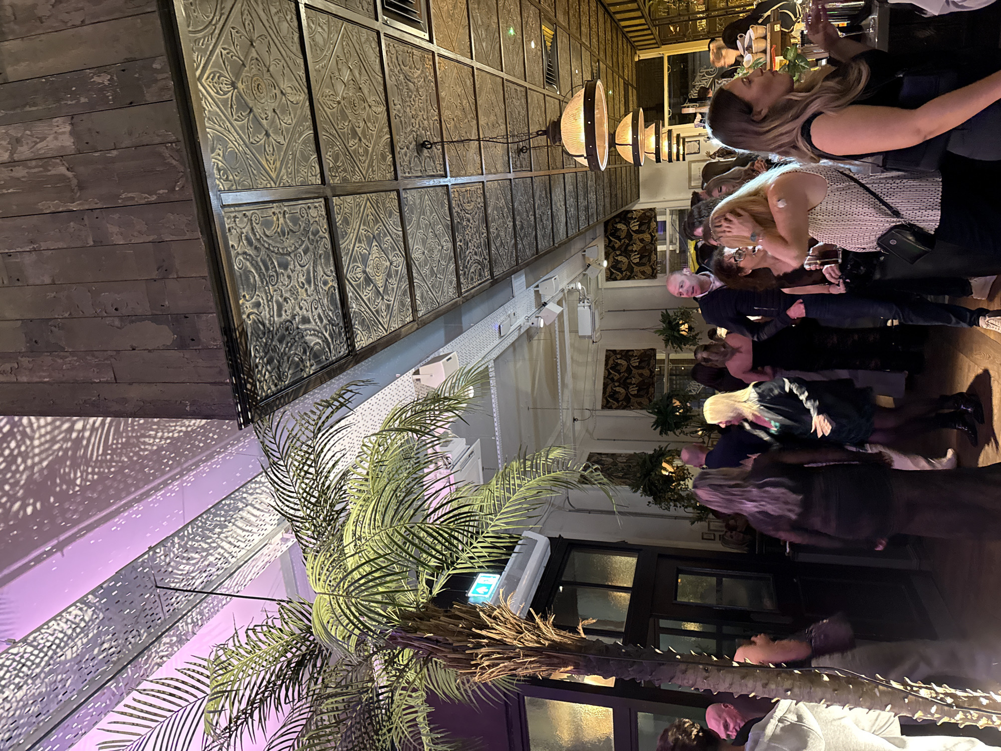 The image depicts a busy indoor scene at an establishment with a distinct architectural style in Ipswich, United Kingdom. A group of people are gathered, some standing and talking, some closer to the bar area where drinks are being consumed. The interior design includes decorative pressed tin panels on the walls, and a palm tree element with lighting casting patterns on the ceiling. The lighting scheme suggests an evening setting. The subjects are dressed in a mix of casual and smart attire. The establishment appears to be a bar or restaurant.