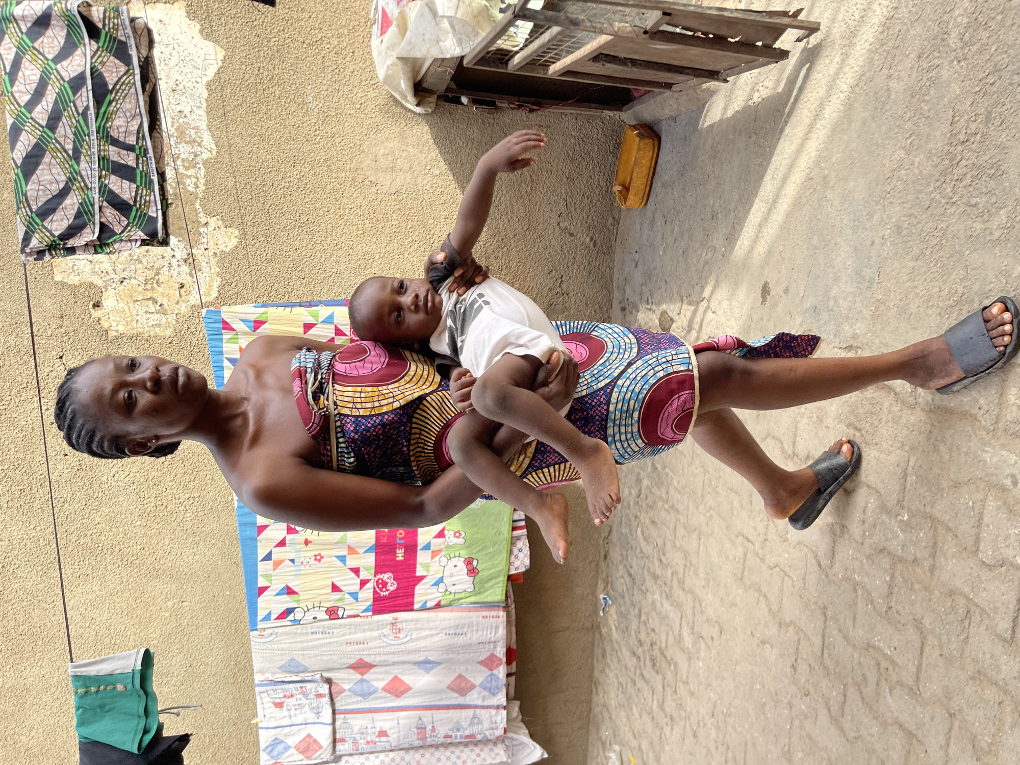 The image shows a woman holding a young child outdoors in bright daylight. The woman has dark skin, braided hair, and wears a colorful, patterned strapless dress or wrapper with circular and geometric designs in shades of blue, pink, and orange. She looks directly at the camera with a neutral expression. Her visible foot is adorned with a dark slider sandal.
She cradles a young child, who also has dark skin and short hair, on her hip or lap. The child wears a white t-shirt and looks towards the camera with a neutral expression, one arm slightly raised.
The background features a textured, light brown wall. In the upper left, a piece of patterned fabric, typical of African prints, hangs on a line. Text visible on this fabric includes "ANKARA MIXED COTTON" and "NIGERIA". Above the child, a wooden structure and a yellow plastic container are visible. The ground is paved with light-colored concrete or interlocking blocks, showing shadows consistent with strong sunlight.
Below the woman and child are neatly folded textiles. One blanket clearly displays a "Hello Kitty" cartoon character, while other fabrics feature geometric patterns and small illustrations of buildings. The word "NUTHAY" is repeatedly printed on a white patterned fabric, and "SAMSUNG" is faintly discernible on another. A small green pouch hangs on a line to the left of the woman's shoulder. The scene depicts a casual outdoor setting in Nyanya, Nigeria.