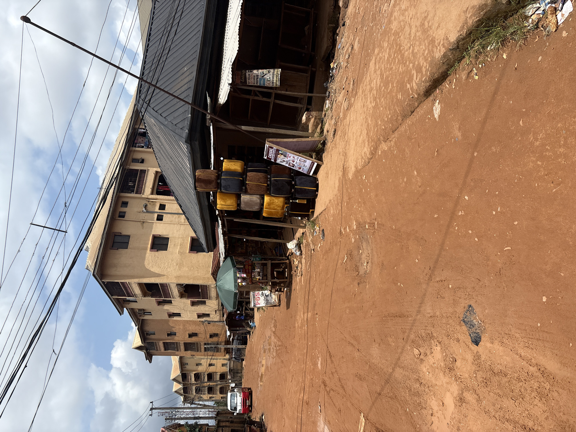 In Nkpor, Nigeria, a street scene unfolds with a red dirt road under a partly cloudy sky. In the background, several multi-story buildings, painted in shades of tan and yellow, line the street. In the mid-ground, a dark corrugated metal roof covers a row of stalls. To the left of these stalls, a green umbrella shades a vendor's table displaying a POS (point of sale) sign. Stacked to the left of the POS table are several upholstered ottomans in brown, gold, and black. To the right of the stalls, a white sign with black text reads "OWBE ENTERPRISE PAY YOUR UTILITY BILLS KILLS HERE PREPAID DSTV GOD." A white and red mini-bus is parked on the left side of the road. The dirt road shows tire tracks and some scattered debris.
