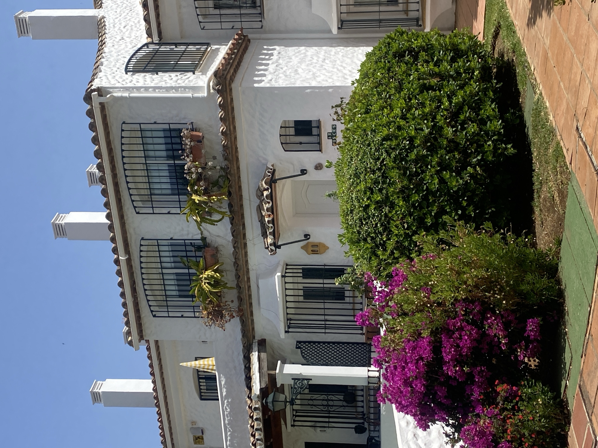 The image shows the exterior of a white, multi-story building with a textured facade and a red tile roof. The building has several windows, many of which are protected by black wrought-iron bars. Some windows have plants or flowers in pots outside them. There are also white chimneys extending from the roof.
In the foreground, there is lush green foliage and vibrant purple bougainvillea flowers, suggesting a warm climate and a well-maintained garden area. To the right, a pathway made of reddish-brown tiles is visible, bordered by grass. The scene is brightly lit, indicating it is daytime, and the clear blue sky suggests fair weather.
There is some visible text on a small plaque on the building near the entrance, reading "32 PISCES". The overall impression is one of a charming, perhaps Mediterranean, residence on a sunny day. There are no people visible in the image.