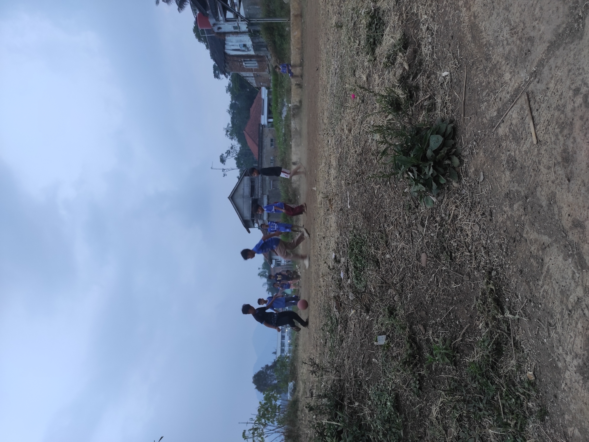 In Sumedang, Indonesia, a group of young boys are engaged in a casual game of soccer on a dusty, uneven field under an overcast sky. The foreground is dominated by dry, sparse vegetation and the dirt ground. In the middle ground, the boys are scattered across the field, with two in the center actively playing with a reddish-brown soccer ball. One boy in a black shirt and pants is kicking the ball, while another in a blue shirt and khaki pants is running towards it. Several other boys are visible in the background, some watching and others walking. The scene is set against a backdrop of rural Indonesian architecture, with simple houses and buildings, some appearing weathered. In the distance, mountains are faintly visible through the hazy atmosphere. The overall impression is one of a simple, everyday activity taking place in a rural Indonesian community.