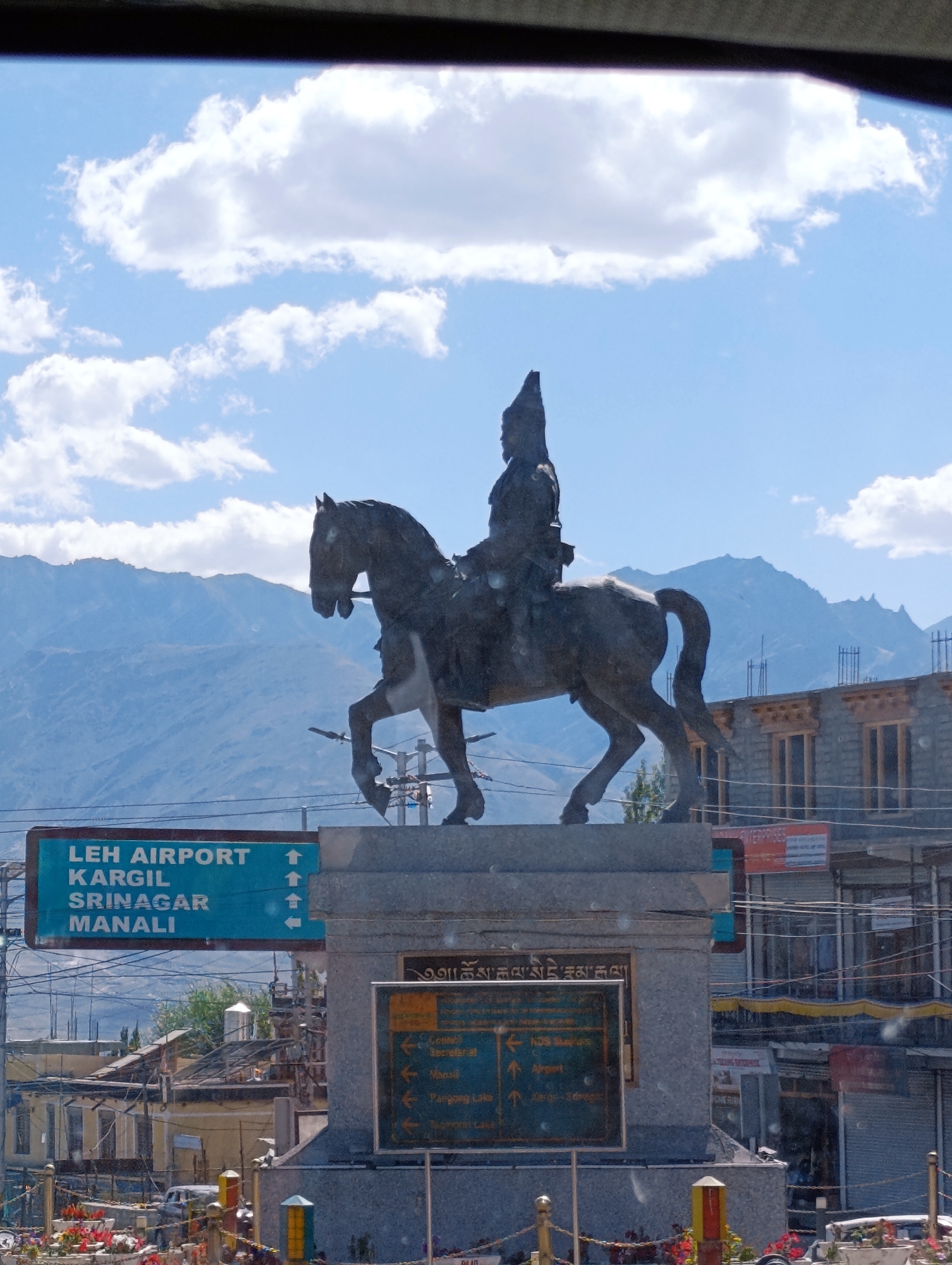 A statue of a man on horseback, cast in dark metal, stands prominently against a backdrop of hazy blue mountains and a sky with scattered white clouds. The statue is positioned on a stone pedestal, which is part of a larger monument. In front of the pedestal, a bright blue sign with white lettering indicates directions to "LEH AIRPORT," "KARGIL," "SRINAGAR," and "MANALI," with arrows pointing upwards. To the right of this, another sign with green lettering lists various locations and directions. Behind the monument and statue, there are buildings with stone facades and wooden window frames, suggesting an urban or semi-urban environment. The overall scene depicts a landmark in Leh, India, likely a memorial or a directional point of interest, with a signpost indicating major routes from the city.