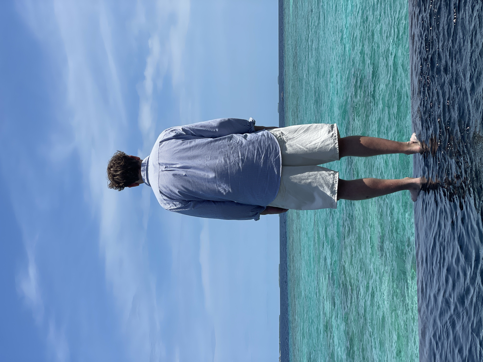 A person stands at the edge of an infinity pool, looking out at the ocean. The person is wearing a blue and white striped shirt and white shorts. The ocean is a vibrant turquoise color, and the sky is a bright blue with scattered clouds. In the distance, a few small islands can be seen. The water in the pool is dark and reflects the sky.