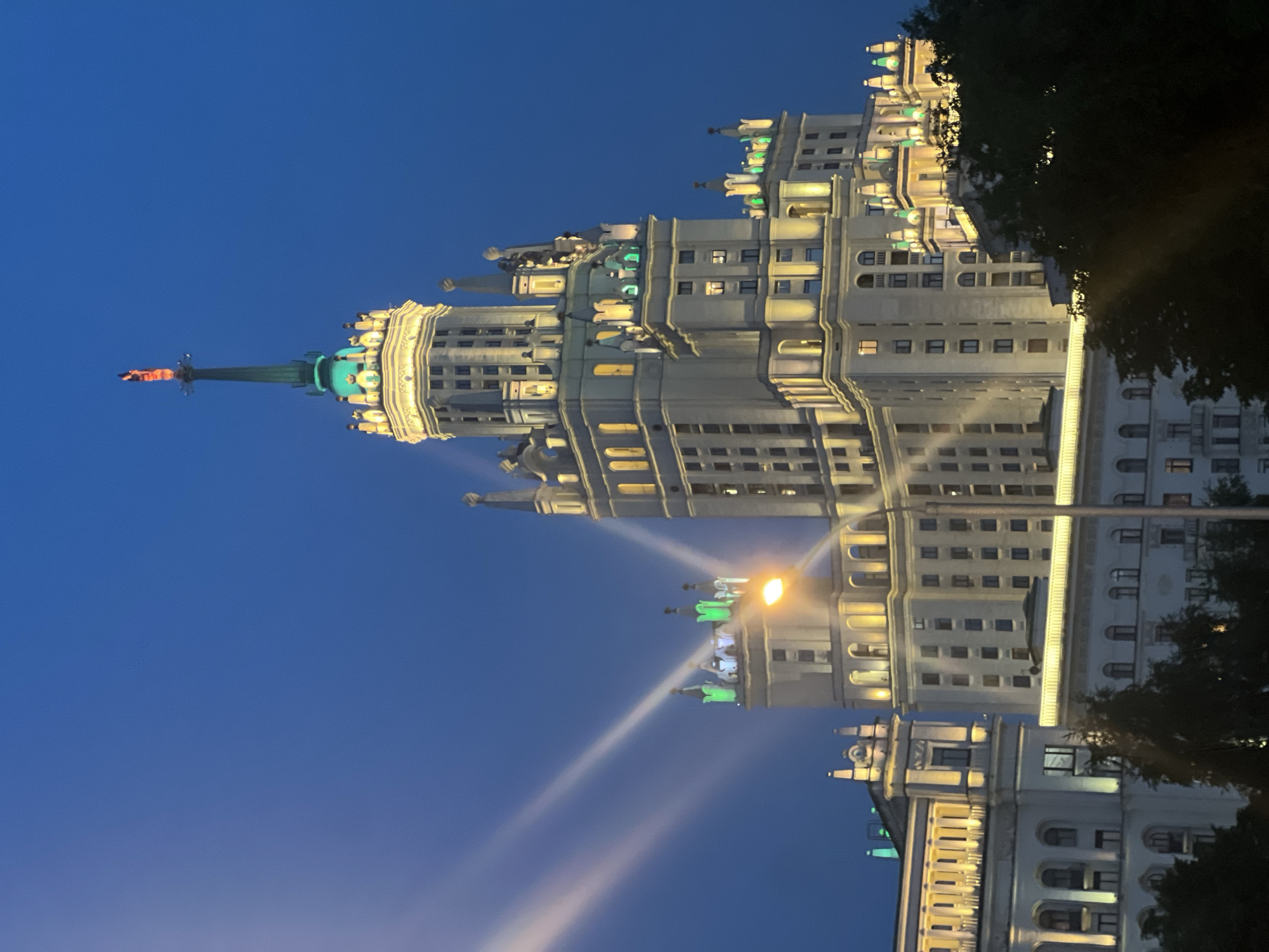 The image shows a view of the Stalinist-era skyscraper, the Kotelnicheskaya Embankment Building, in Moscow, Russia, at dusk. The building's architecture is characterized by its multi-tiered structure and ornate spires. The skyscraper is illuminated with yellow and green lights, highlighting its details. A statue is visible at the very top of the building's spire. The sky is a deep blue, indicative of twilight. In the foreground, there are trees, and a streetlamp is also visible, casting a warm glow. There are no people visible in the image.