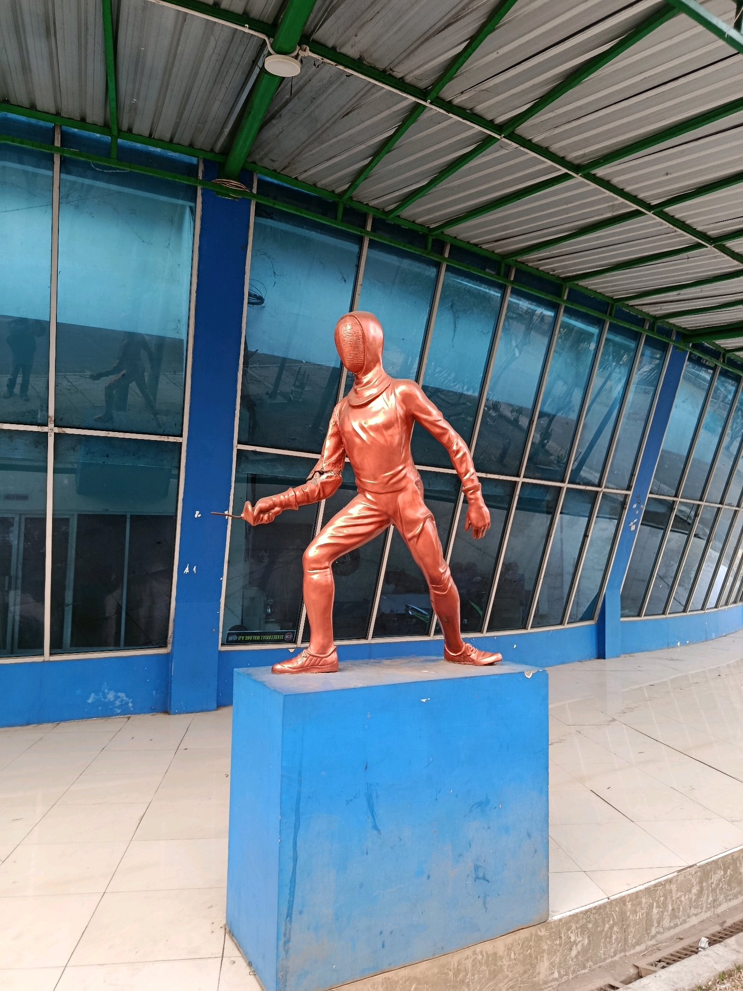 A bronze statue of a fencer stands on a blue pedestal in front of a building with large, slanted blue windows. The fencer is in a lunging pose, holding a foil. The building appears to be a modern structure with a metal roof supported by green beams. The ground in front of the building is paved with light-colored tiles. The scene is outdoors, likely in a public area. The location is identified as a part of the Velodrome outdoor sports complex in Malang, Indonesia.
