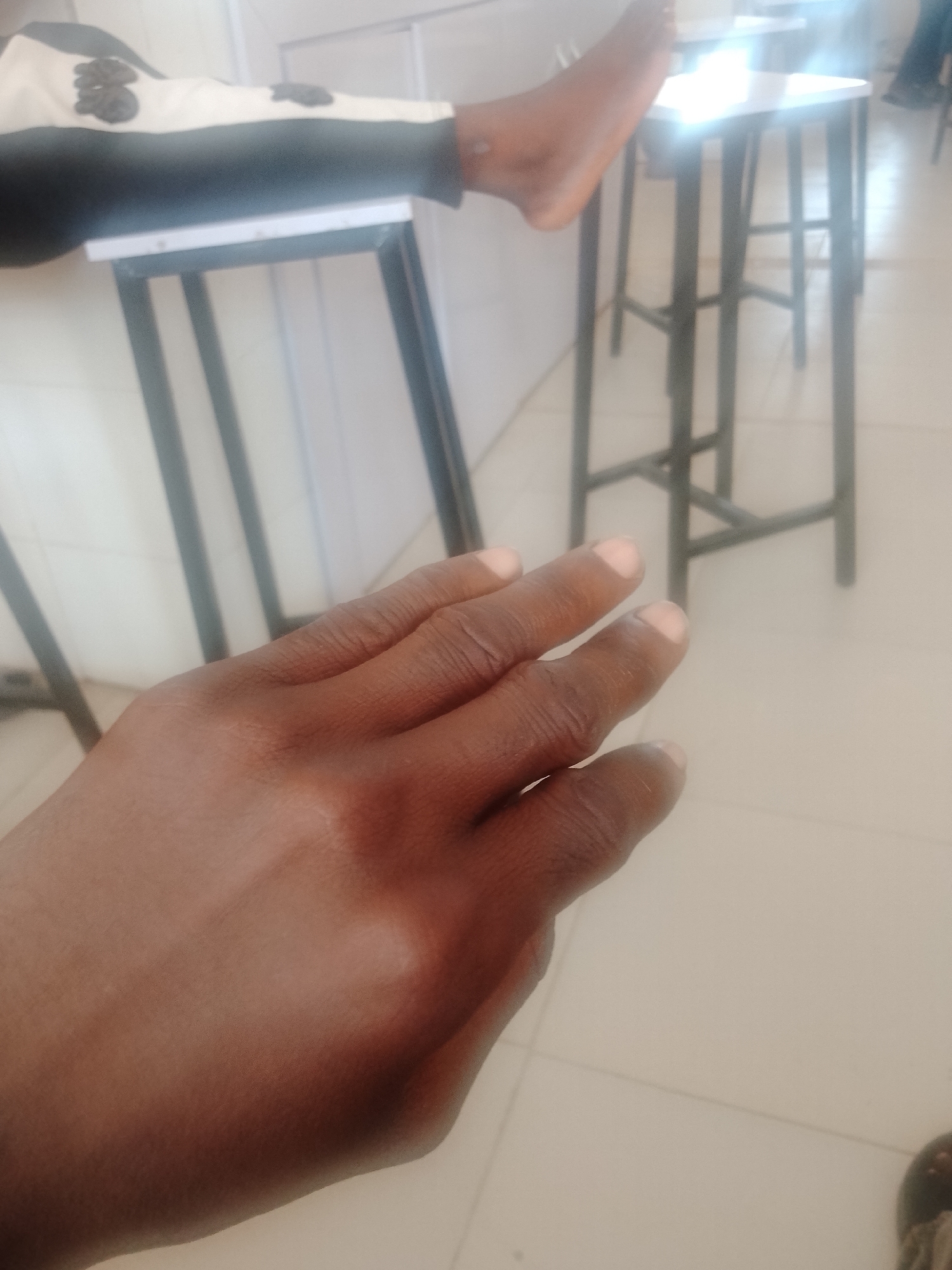 In an indoor environment with a tiled floor, a person's left hand is prominently featured in the foreground, appearing to be resting on a surface. The hand is dark-skinned and shows the knuckles and portions of the fingers. In the background, slightly out of focus, are several tall, stool-like objects with dark metal frames and light-colored tops. Further in the background, a person's lower legs and feet are visible, one leg resting on a flat surface. The clothing on the visible leg is light-colored with dark embellishments. The scene suggests a casual or possibly educational setting, with the furniture indicating a communal space like a classroom or waiting area.