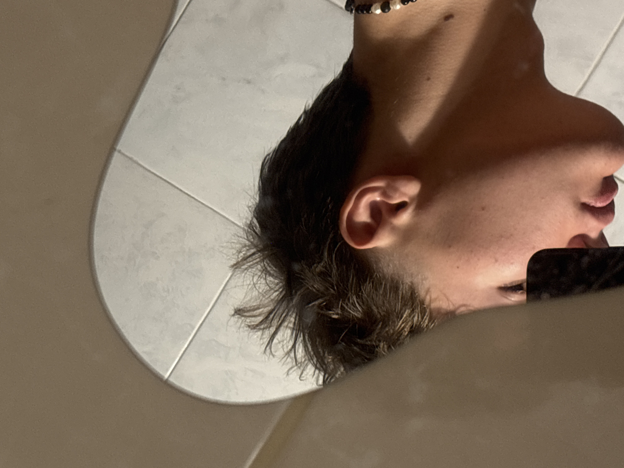 The scene is an interior location in the city of Troyan, Bulgaria. The image depicts the reflection of a young individual, seen in left profile, within an irregularly shaped mirror. The individual has short, dark brown hair with a textured, somewhat spiky style on top and shorter sides. Their right ear, jawline, chin, and closed lips are visible; the right eye is partially obscured by a dark, rectangular object held near the face, consistent with a smartphone. A necklace composed of alternating black and white/cream beads is worn around the individual's neck. The background reflected within the mirror consists of light-colored, rectangular or square tiles with light gray grout lines. The surface surrounding the mirror's reflection is a solid, dull brown or beige color. Lighting is ambient, creating subtle shadows beneath the chin and along the neck.