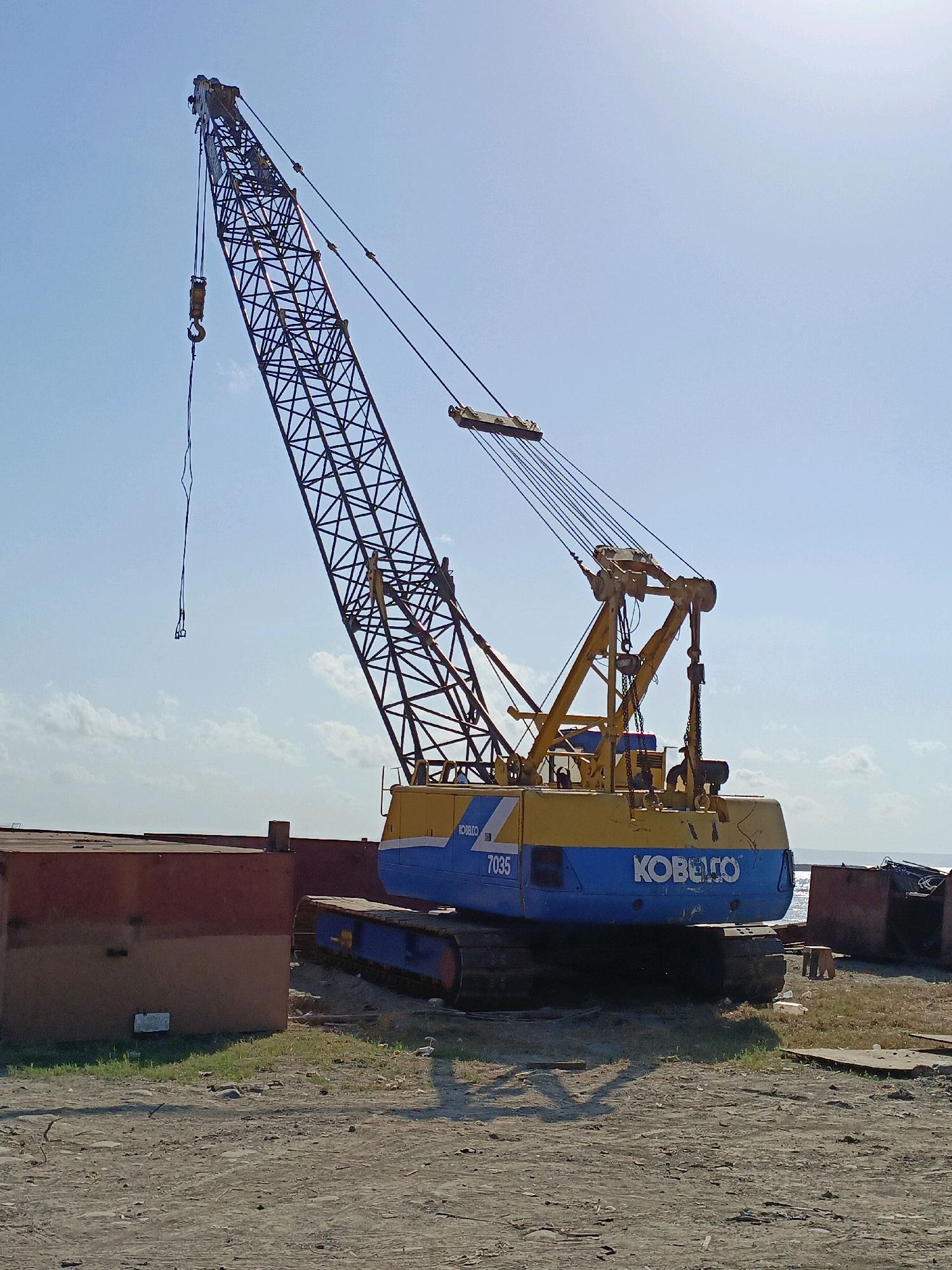 A large, yellow and blue Kobelco 7035 crawler crane is parked on a dirt lot near the water's edge, likely in Bulukumba, Indonesia. The crane's boom is extended upwards and slightly to the left, with a hook hanging from it. The crane's body is blue with yellow accents and the Kobelco logo is visible. In the foreground, the ground is covered in dirt and scattered debris. To the left and right of the crane are large, reddish-brown metal structures, possibly part of a shipping container or barge. The sky is clear and blue with some scattered clouds, and the sun is shining brightly, casting shadows on the ground. The environment appears to be an industrial or shipyard area, with the presence of the crane and the large metal structures suggesting ongoing construction or loading/unloading operations. There are no people visible in the image.