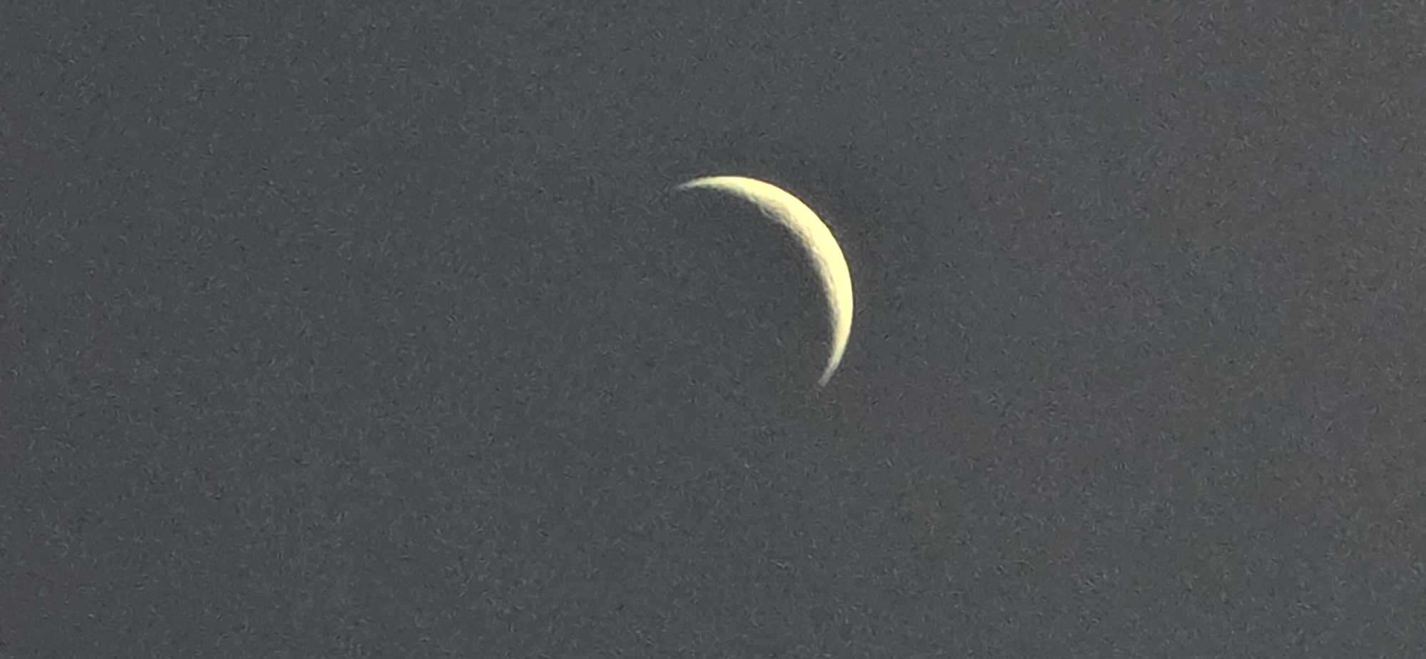 A crescent moon is centered against a dark gray sky. The location in Benasque, Spain, is not discernible from the image.
