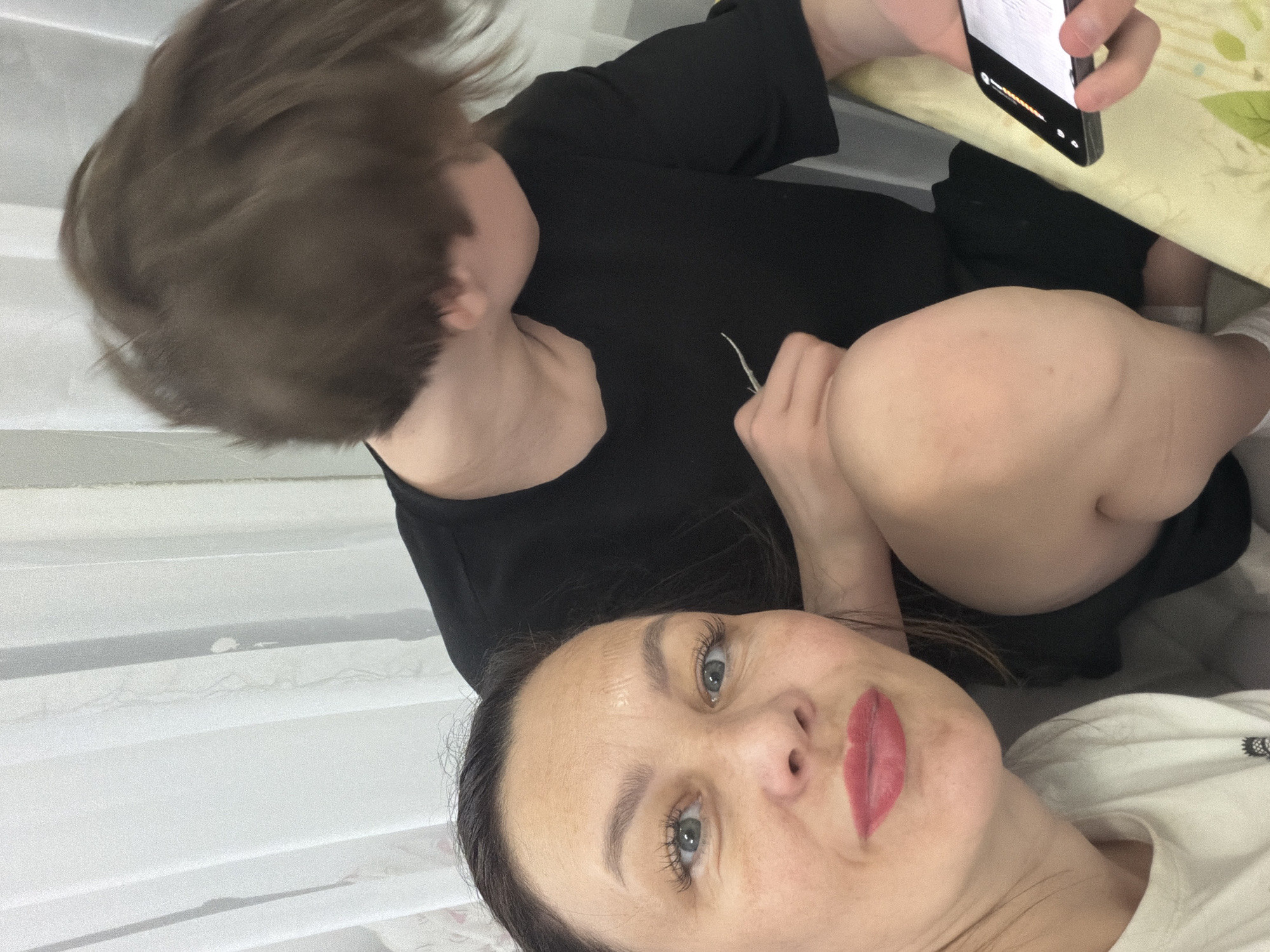 The image captures a candid indoor moment featuring a woman taking a selfie with a child nearby. The woman, with dark hair, blue eyes, and red lipstick, is positioned in the foreground, looking directly at the camera with a neutral expression. She is wearing a white t-shirt.
Behind and slightly above her, a young child, possibly a boy, is visible with short brown hair. The child's face is turned away from the camera, and they are wearing a black top. In the child's hand, or held by someone close to the child but out of frame, is a black smartphone. The phone's screen shows some interface elements, including what appears to be a progress bar or status indicators, but no discernible text. The phone is positioned over a light green or yellowish table surface with a subtle pattern.
The background consists of a white, possibly textured or patterned wall covering, or perhaps vertical blinds, suggesting an indoor home setting. The overall lighting is soft, indicating an indoor environment rather than bright natural sunlight. The image appears to be a casual snapshot taken in Chaikovsky, Russia.