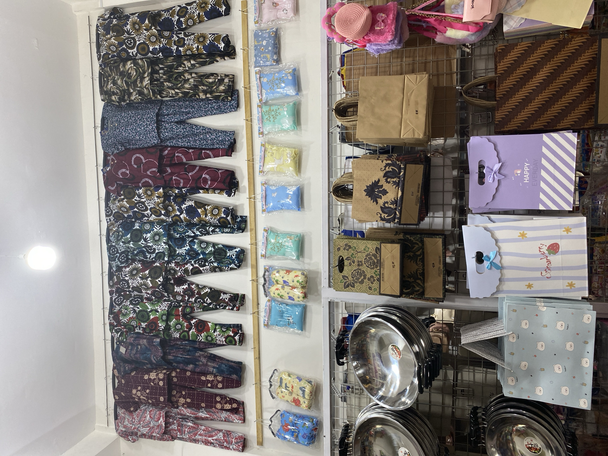 In a retail store located in Lamteumen Barat, Indonesia, a wall-mounted display showcases a collection of clothing and accessories. The upper section features rows of patterned pajamas, hung on individual hangers against a white wall. Below, a horizontal shelf holds several clear plastic bags containing folded items, possibly infant clothes or accessories. Further down, on metal shelving units, various items are displayed. On the left, stacks of shiny, silver metal bowls or woks are arranged. To their right, a collection of paper gift bags in different designs and colors are visible. Some of these bags have handles and feature floral patterns, striped designs, or cartoonish characters like bears. A small pink purse with a circular straw hat embellishment is also hanging on the right side of the display. The store appears to be well-lit, with a single light fixture visible at the top of the frame.