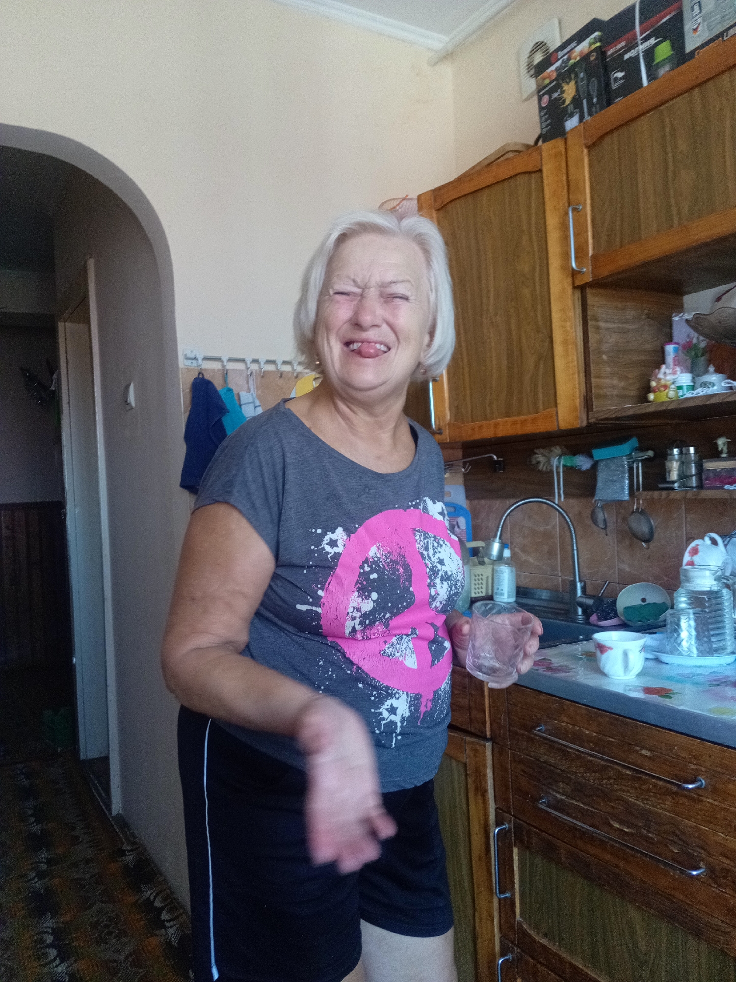 In a domestic setting, likely a kitchen, an elderly Caucasian woman with short, white hair is playfully sticking her tongue out at the camera. She is wearing a dark gray t-shirt with a large pink and white graphic and black athletic shorts. She is holding a clear glass in her right hand, and her left arm is bent, with her hand blurred due to motion. The background shows wooden cabinets, a kitchen sink with a faucet, and various household items on shelves and countertops. The overall lighting suggests an indoor environment. The image does not contain specific landmarks or clues that would definitively place it in Tiachiv, Ukraine.