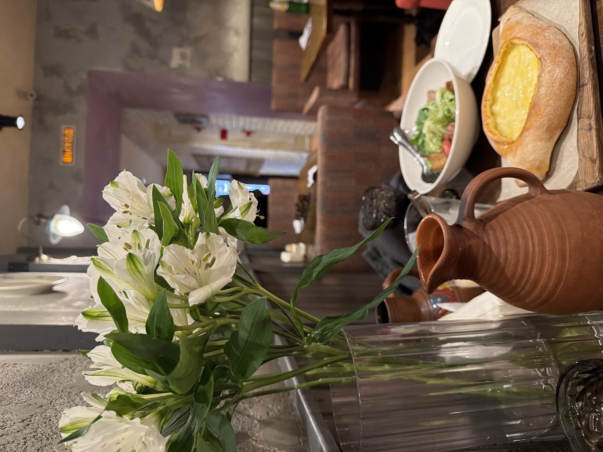 A slightly blurry, medium-shot image presents a culinary scene, possibly in Saint Petersburg, Russia. A bouquet of white alstroemeria flowers in a transparent vase occupies the lower left foreground.
To the right of the flowers is a setting of food, including a brown ceramic pitcher, a white bowl of salad, and a khachapuri (Georgian cheese bread) on a wooden board. The setting implies a restaurant or dining environment.
The background features interior architecture with brick elements, and through an archway, there's a glimpse of a more distant space, indicating a multi-room establishment. The lighting is moderate, suggesting an indoor setting with artificial illumination. No people are visible in the shot.