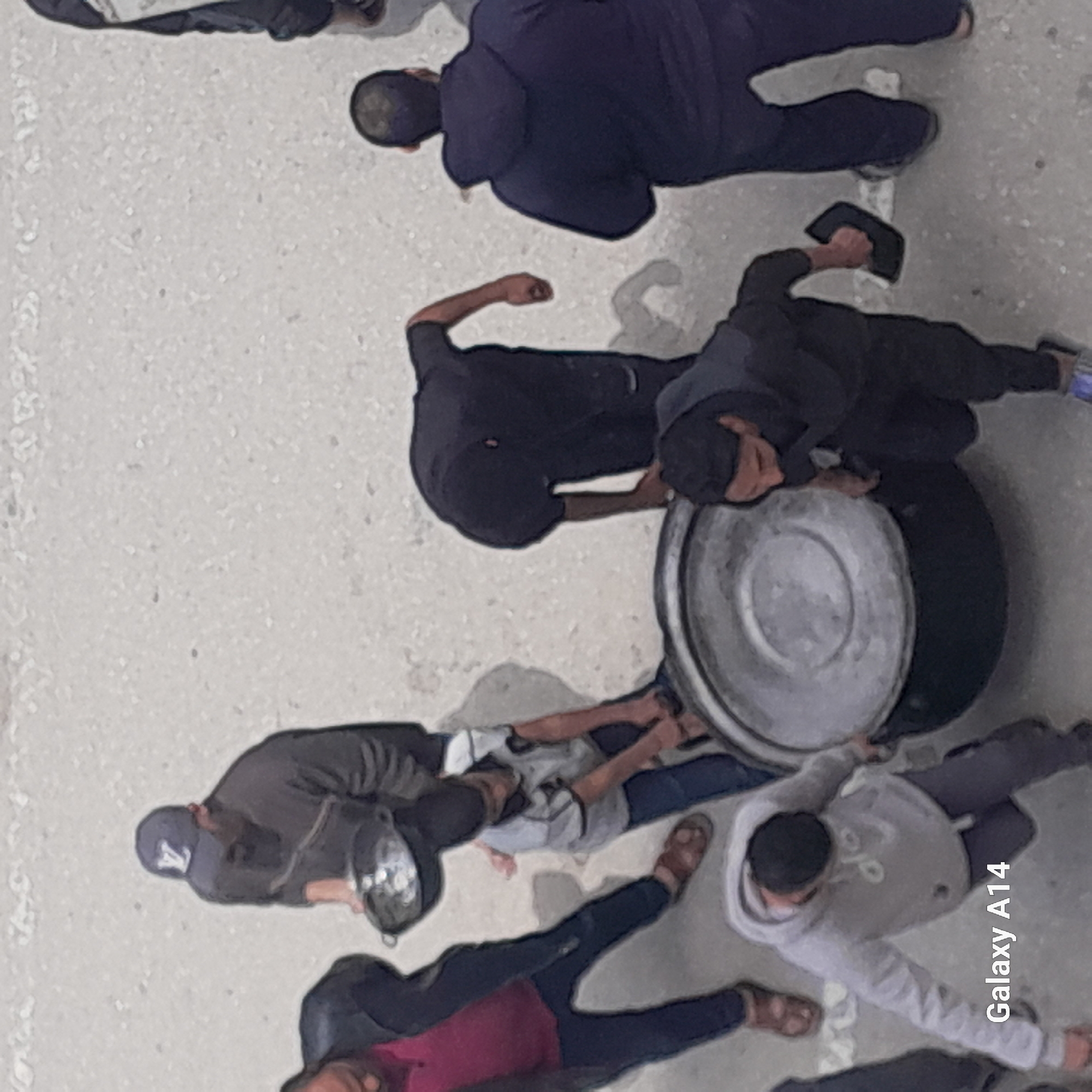 The image depicts a group of people congregated around a large metal drum in an outdoor setting. The environment appears to be a paved area, possibly a street or courtyard. The people are dressed in casual clothing. Their activities and interactions are difficult to determine from the image. No particular objects or subjects besides the people and the drum are readily identifiable. The location of the city is not specified.