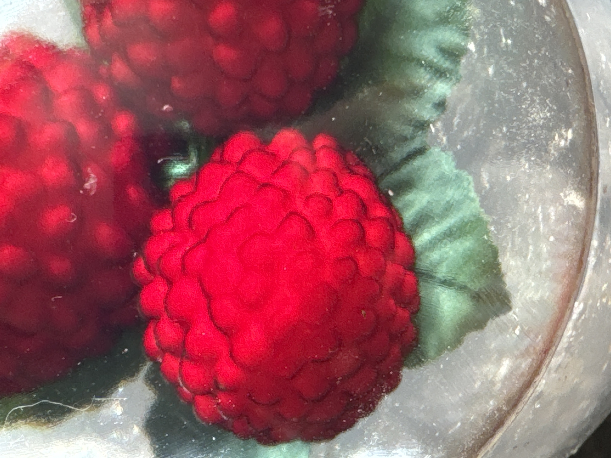 This is a close-up, overhead, still photograph of three raspberries, two in focus and one slightly out of focus, against a muted, greyish background that appears to be the surface of a dish or platter. The raspberries are a vibrant red, with visible drupelets that give them their characteristic textured appearance. Green leaves are visible beneath and between the raspberries, suggesting they are part of a decorative arrangement. The overall lighting is soft, highlighting the glossy texture of the fruit. The image does not depict any identifiable location within the city of Waterford, Ireland.