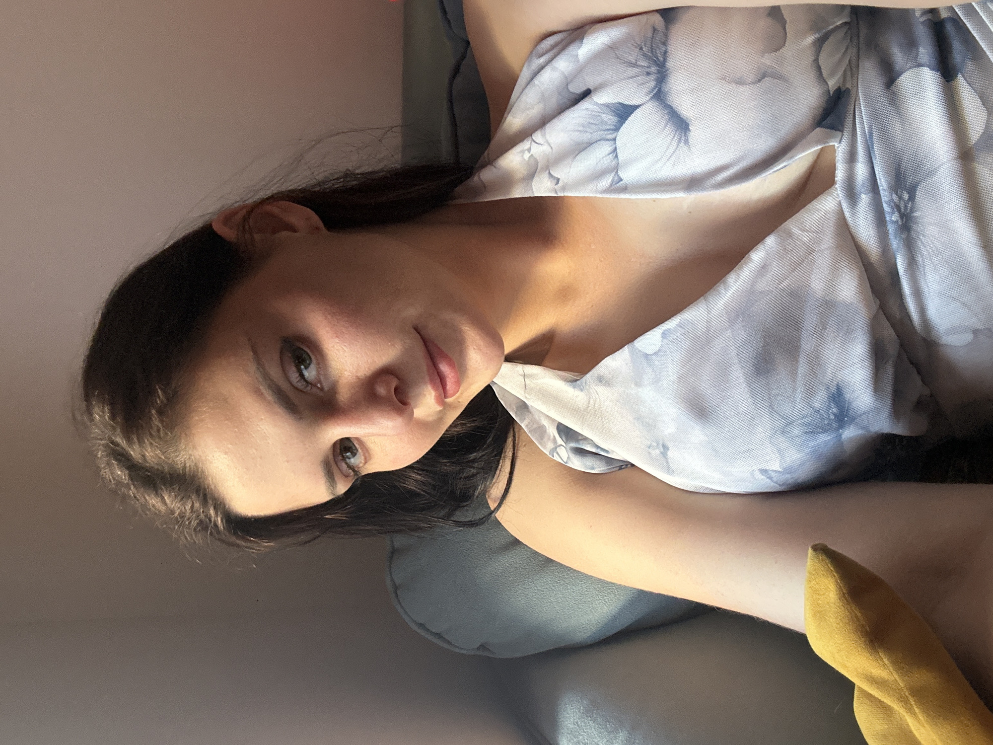 A woman with dark hair and green eyes is sitting on a blue couch, looking at the camera with a slight smile. She is wearing a light-colored dress with a floral pattern. A yellow object is visible on the left side of the image. The background is a wall, with sunlight casting shadows on the couch. The image appears to be taken indoors.