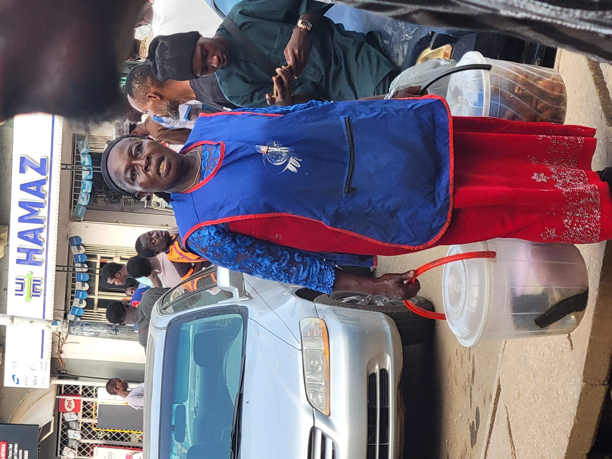 A woman in a red dress and blue apron stands on a street in Jos, Nigeria, holding a bucket with a dark object inside. She appears to be interacting with people near a silver car. Behind her, a group of people are gathered outside a shop with "HAMAZ" written on the storefront. Another man stands nearby, using a mobile phone. The setting is a bustling street scene with shops and pedestrians.