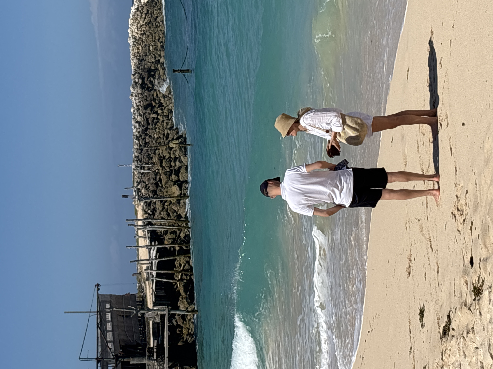 The image depicts a serene beach scene with two individuals standing near the water's edge. A man, wearing a white t-shirt and black shorts, faces away from the camera, looking out at the ocean. Beside him, a woman in a white shirt and a straw hat also faces away, holding a dark object, possibly a phone, in her hand. She carries a light-colored straw bag over her shoulder. The sun casts a distinct shadow of the woman on the sandy shore, suggesting it is a sunny day, likely late afternoon or early morning. The turquoise water gently laps at the shore, with a rocky structure and a wooden pier visible in the background, hinting at a coastal location. The atmosphere appears peaceful and relaxed. There is no visible text in the image.