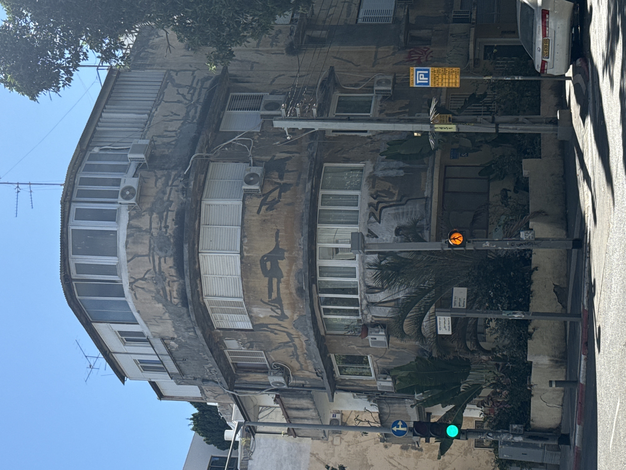 The image shows a corner building with a weathered, discolored facade, likely in Tel Aviv, Israel. The building has multiple stories with a rounded corner on the upper floors featuring large windows. Various air conditioning units are mounted on the exterior walls. The facade is adorned with what appears to be black graffiti or paint streaks. In the foreground, a traffic light displays a green signal. Across the street, a pedestrian crossing signal shows an orange walking person. A yellow parking sign with blue lettering is visible on a pole. A white car is parked on the right side of the street. Lush green plants, including palm leaves, are growing around the base of the building. The sky is clear and blue.