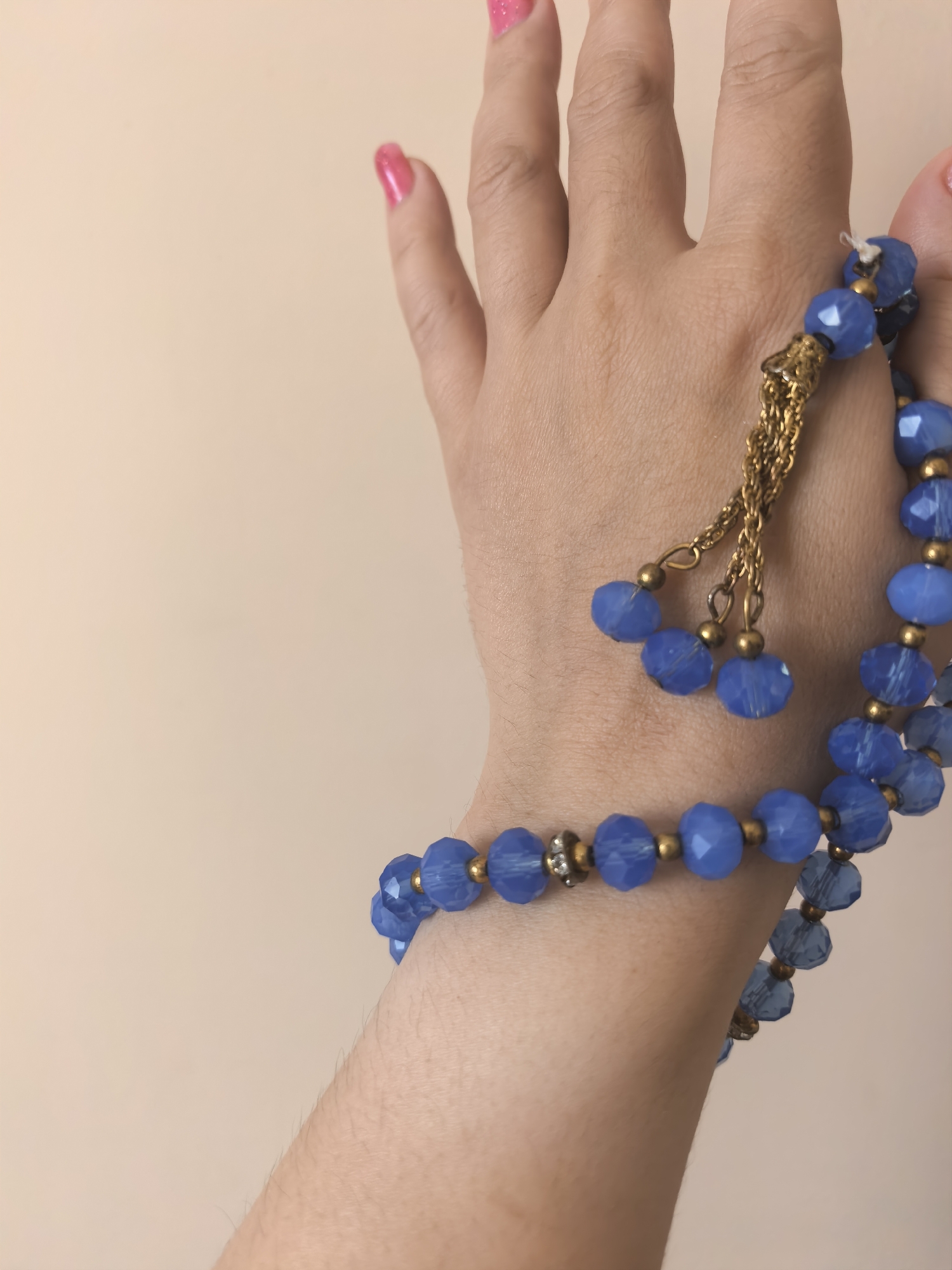 This media file features a close-up shot of a person's hand adorned with a blue beaded bracelet. The bracelet is made of faceted, translucent blue beads interspersed with smaller gold-colored beads. A decorative gold chain dangles from the bracelet, featuring three smaller blue beads at its end. The person's fingernails are painted a vibrant pink. The background is a plain, soft peach or cream color, suggesting an indoor setting with diffused lighting. There is no visible text. The image focuses solely on the hand and the jewelry, with no other context provided.