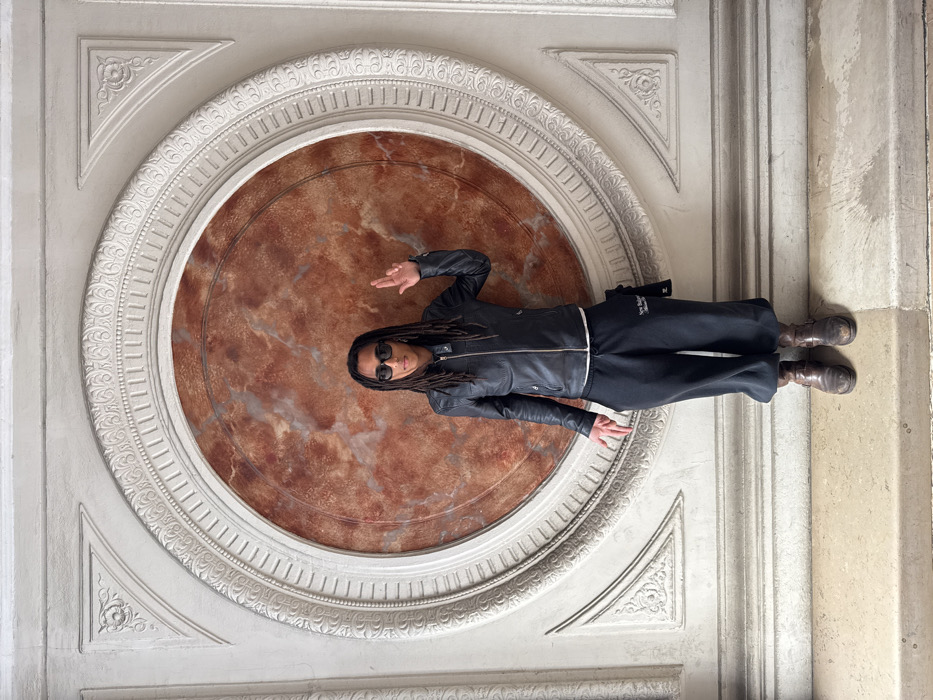 A person with dark skin and dreadlocks, wearing black sunglasses, a black leather jacket, black sweatpants with "New Balance" branding, and dark brown boots, stands centered against a highly ornate wall. The individual's right hand is raised, palm facing forward with fingers splayed, while the left hand is lowered, also with fingers splayed. The wall behind features a large circular medallion filled with reddish-brown, mottled material, likely marble, encircled by multiple concentric rings of intricately carved white plaster or stone. This decorative frame includes classical motifs such as fluting and floral patterns. The floor beneath the individual is composed of light-colored, segmented stone. This architectural setting, characterized by the ornate classical detailing and circular marble inlay, is located within the Louvre Museum in Paris, France.