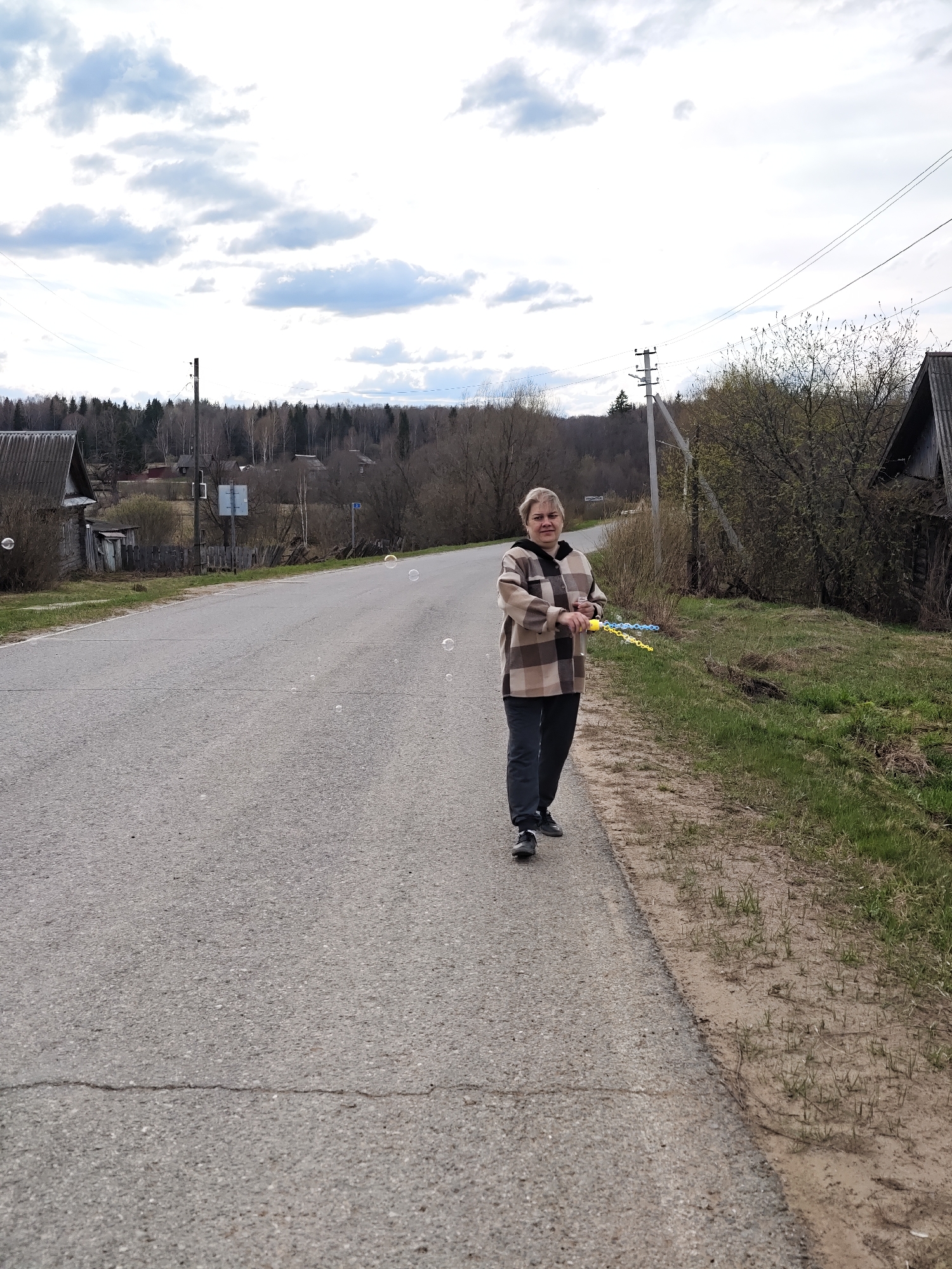 A middle-aged woman stands on the asphalt edge of a rural road in Уральское, Russia. She wears dark pants and a tan and brown plaid jacket, holding a yellow and blue toy. Several soap bubbles float in the air around her. To her left, a weathered wooden building sits near the road. Behind her, a grassy area slopes down to a line of trees and a cloudy sky. Power lines stretch across the top of the image. A road sign is visible in the background near some trees.
