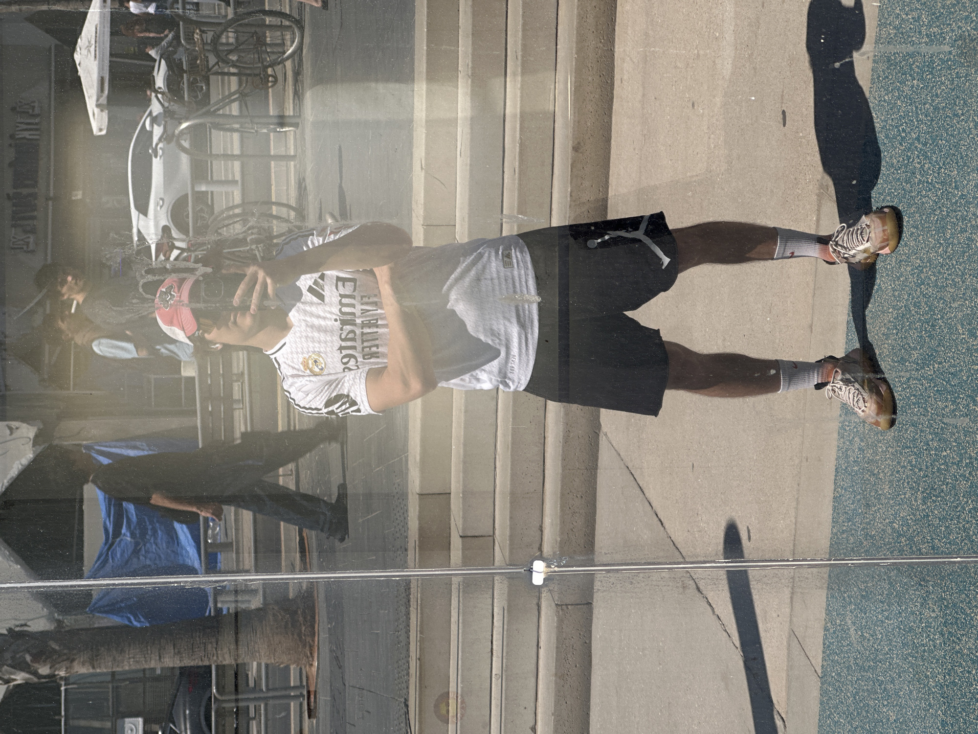 The image displays a complex reflection on a large glass surface, likely a shop window or building facade, in an urban setting. The central subject is a young man reflected prominently, wearing a white Real Madrid football jersey. The shirt features a mirrored Adidas logo and the mirrored text "SƎTAЯIMƎ" ("Emirates"), along with the mirrored Real Madrid crest. He also wears black shorts with a white Jordan logo, white socks, light-colored sneakers, and a red and white cap. He is holding a smartphone or camera, appearing to take a photo or video.
The ground visible in the reflection comprises a light grey concrete sidewalk alongside a distinct blue textured surface. The man's long, dark shadow is cast across this ground, indicating bright, strong sunlight. In the background reflections, a white car, several bicycles (some on a bike rack), and concrete steps are discernible. A palm tree trunk is also visible in the lower left section of the reflection, along with other indistinct figures and urban elements.
The scene takes place on a clear, sunny day, with the long shadows suggesting late morning or early afternoon. The urban environment, featuring buildings, vehicles, bicycles, and a palm tree, aligns with the context of a city like Tel Aviv. The image's reflective quality overlays various visual elements, creating a dynamic portrayal of the street life.