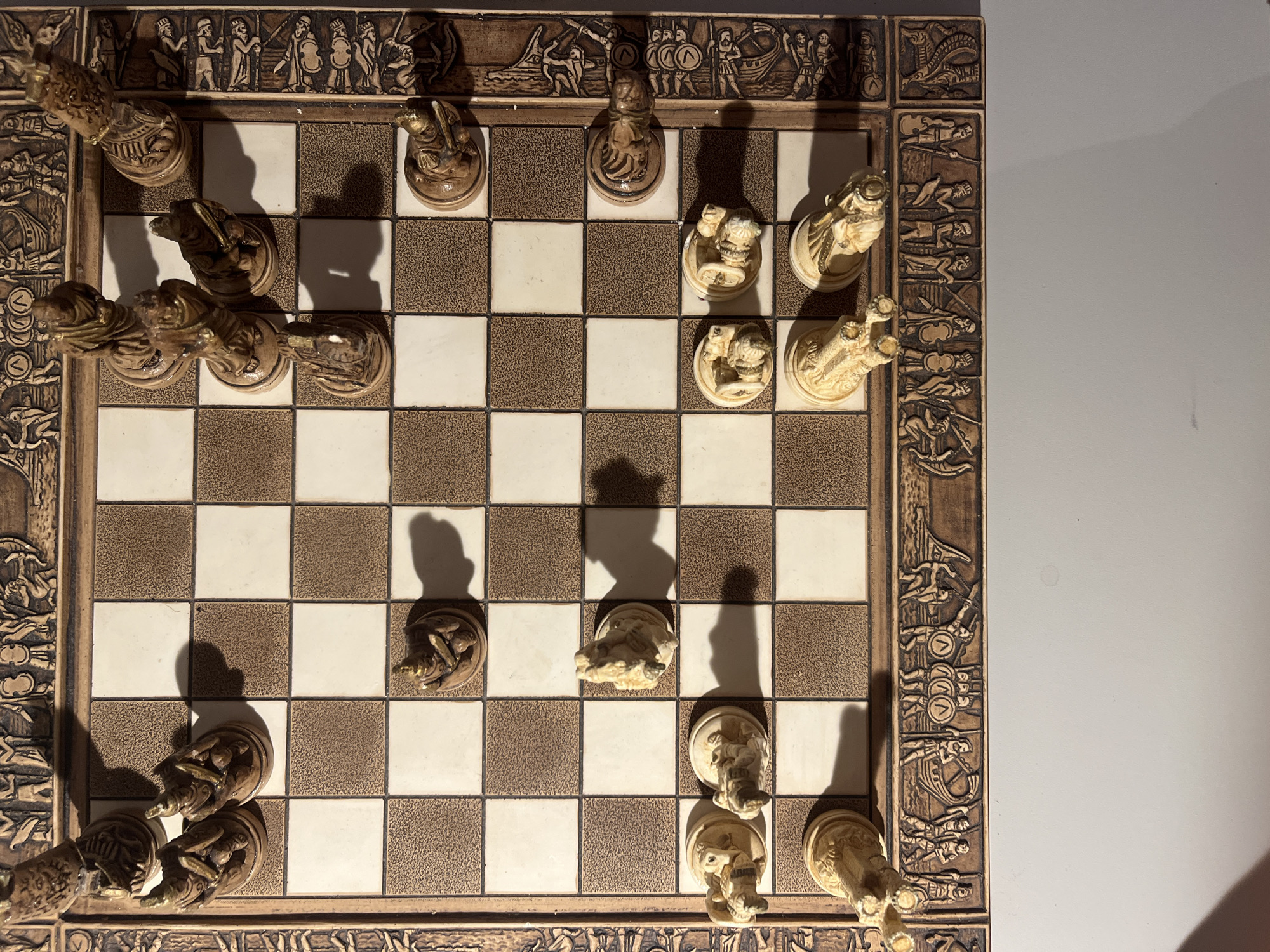 The image shows a close-up, overhead view of a wooden chess board with intricately carved borders depicting ancient scenes. The chess pieces are stylized, made of a light-colored material with gold accents, and appear to be of an ancient design, possibly Egyptian or Roman. The chess board itself has alternating light and dark brown squares. Several chess pieces are scattered across the board, suggesting a game in progress or recently ended. The lighting creates distinct shadows from the pieces, indicating a strong light source from the top left. The background is a plain, light-colored wall. There is no indication of location within Minsk, Belarus, or any other specific geographical context.