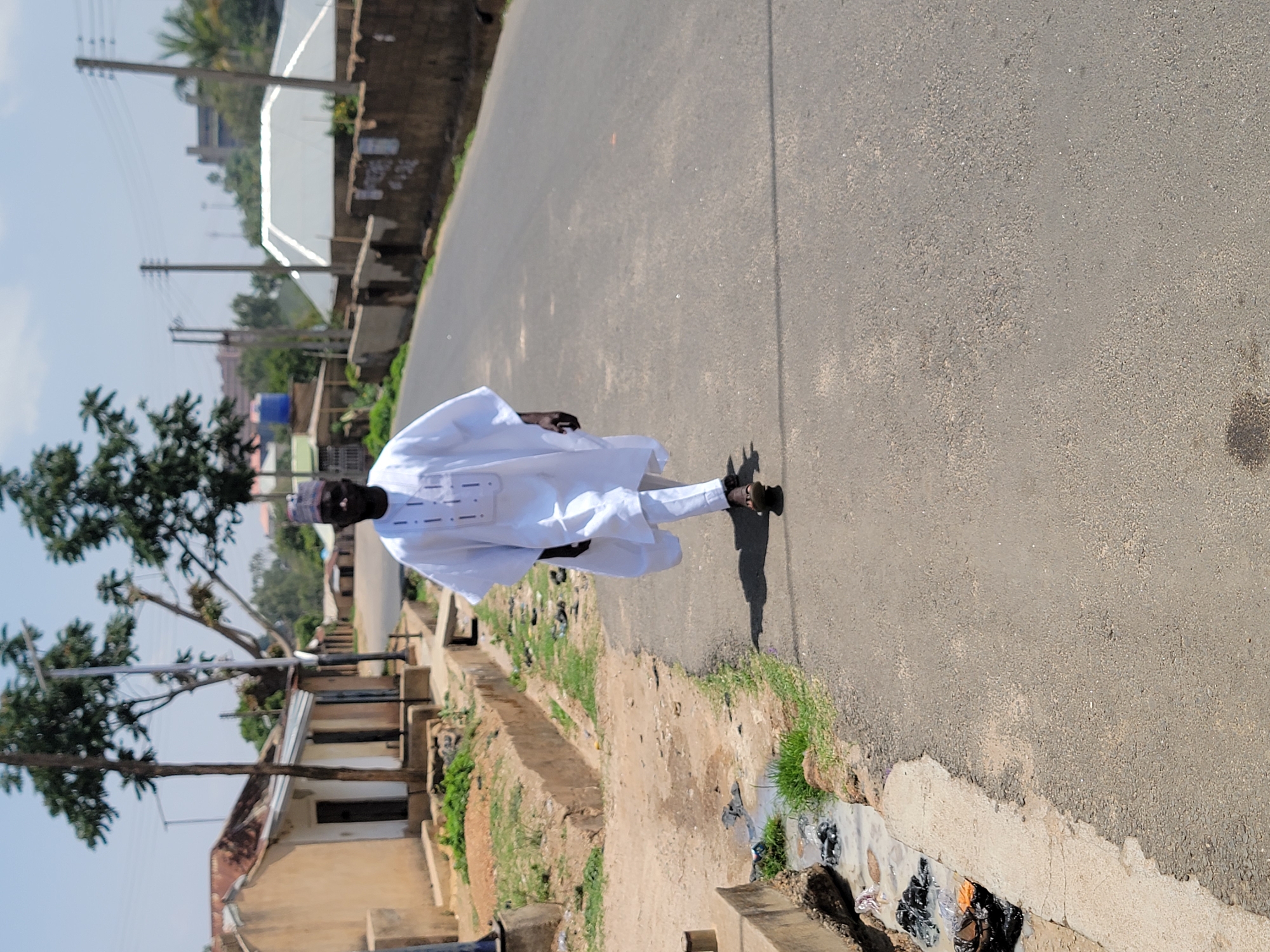 The image shows a man wearing a white traditional garment walking on a paved road. The man is dressed in a white kaftan and trousers. He is wearing a cap. The road runs through a residential area with buildings and trees visible on either side. Along the side of the road is a drainage ditch that has some trash in it. The location may be Jos, Nigeria.