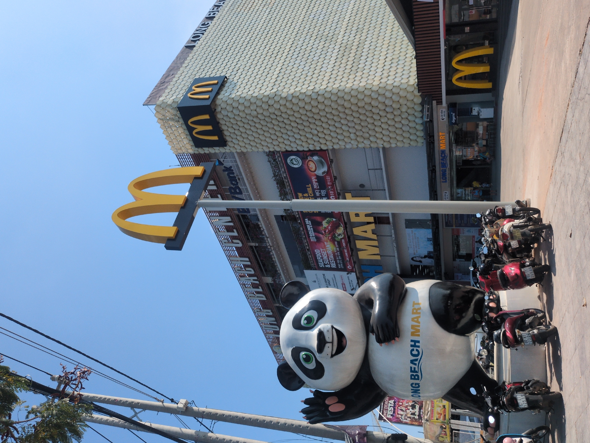 A scene in Phú Quốc, Vietnam, shows a McDonald's establishment with unique architecture. The building features a façade adorned with numerous white discs, resembling scales. The McDonald's logo is displayed in yellow and black, on the roof and prominently on a sign. In front of the building stands a large panda statue advertising Long Beach Mart. Several motorbikes are parked along the street. The sky is clear blue. Power lines run across the lower left corner of the frame.
