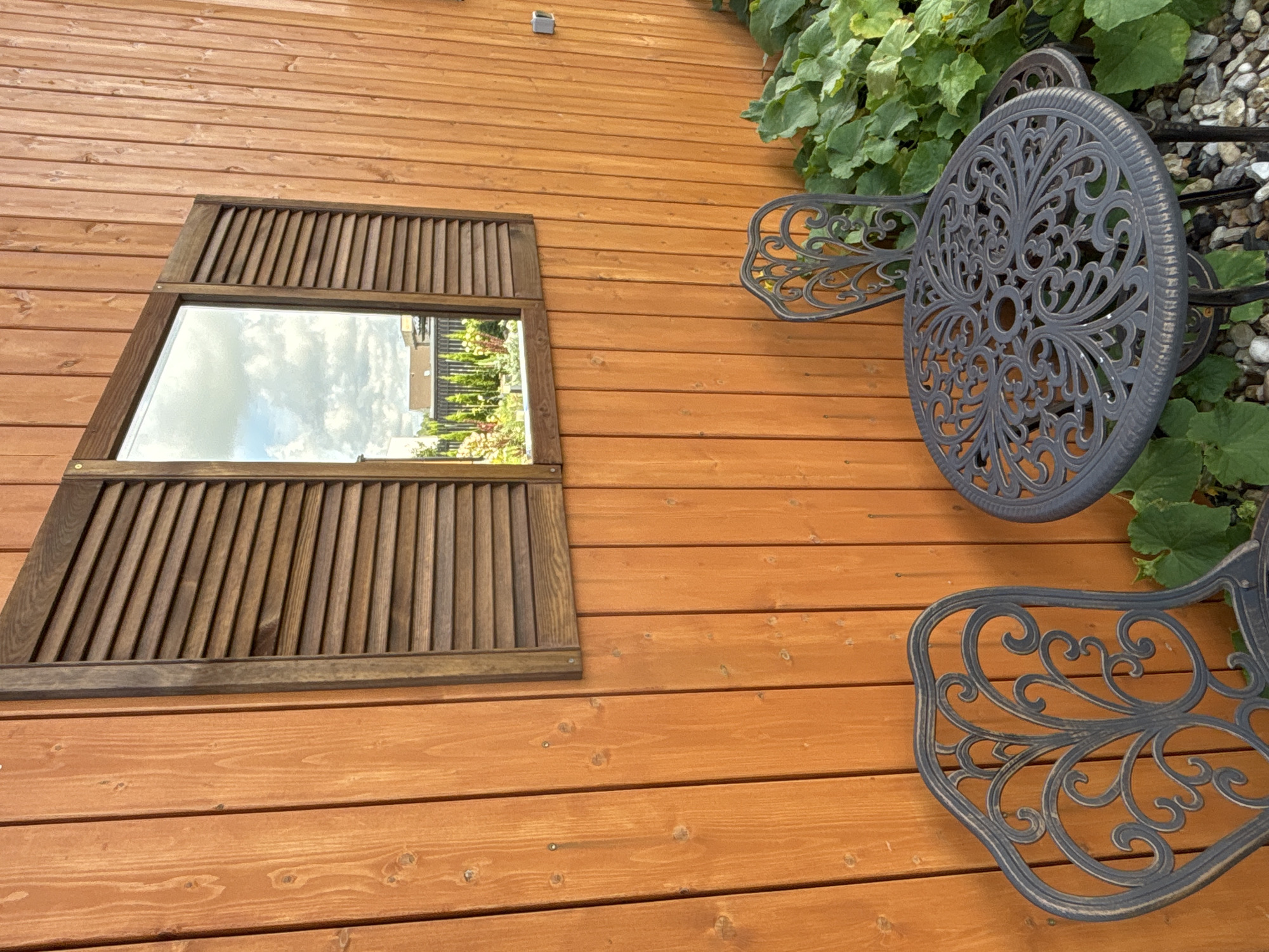 An outdoor scene, possibly in a garden area of Zvenigorod, Russia, features a wooden wall with a louvered shutter and mirror. In front of the wall, a round, ornate metal table and two matching chairs are set up. Lush green foliage grows around the furniture. The mirror reflects a cloudy sky and a distant garden.