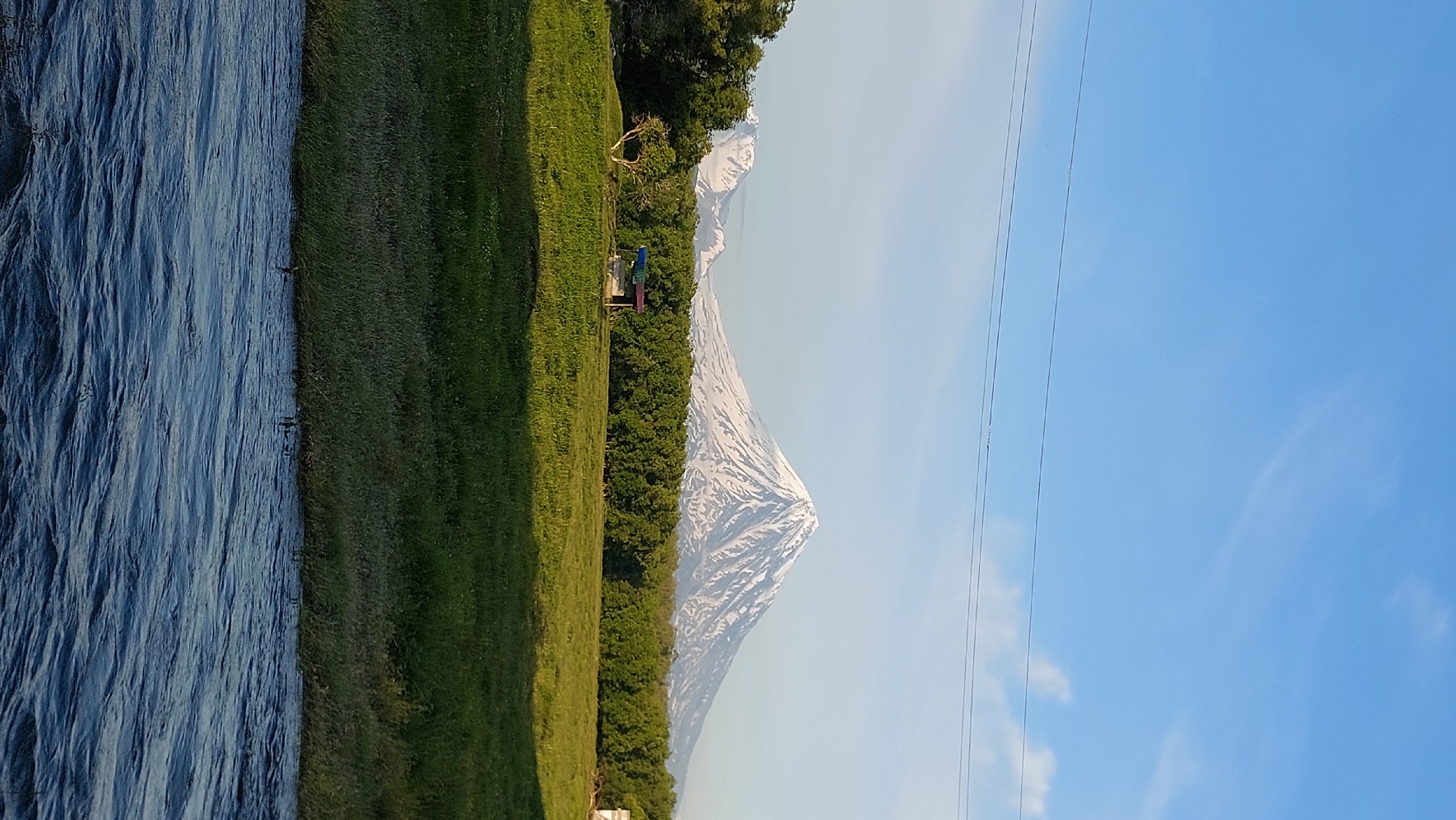 The image depicts a serene natural landscape featuring snow-capped mountains in the background, likely part of the volcanic chain in the Kamchatka Peninsula, Russia. In the mid-ground, a dense line of lush green trees forms a natural barrier, with a small, rustic-looking shelter or shed situated in a clearing among them. A solitary, gnarled tree stands to the left of the shelter. The foreground is dominated by a vibrant green grassy field, gently sloping down towards a body of dark blue water, possibly a river or lake, with visible ripples on its surface. The sky above is a clear, bright blue, with faint wisps of clouds and two parallel power lines stretching across the upper portion of the frame. The overall impression is one of tranquility and natural beauty, characteristic of a rural or wilderness setting.