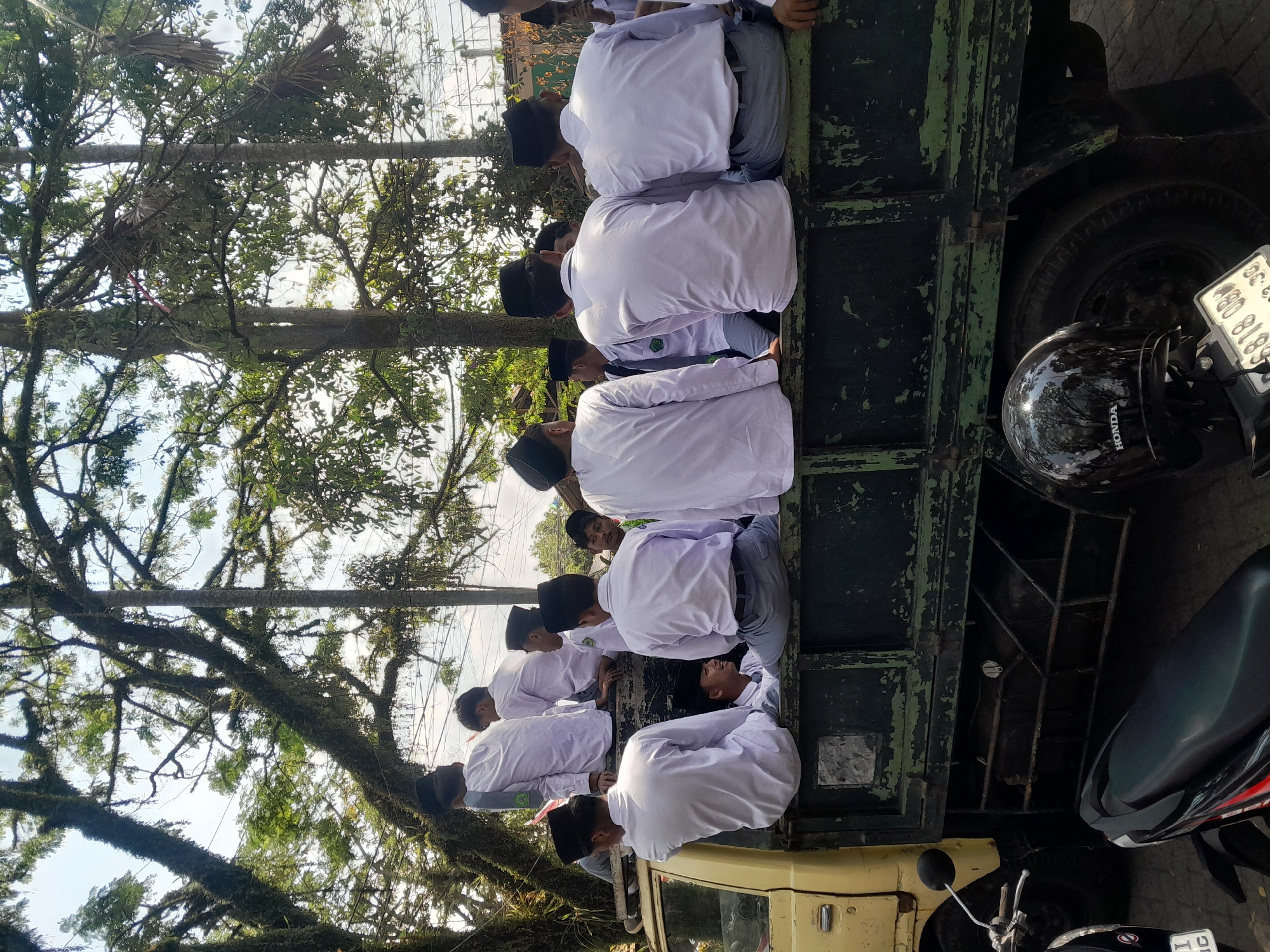 In Genteng, Indonesia, a group of young males, presumably students wearing white uniforms and black peci hats, are seated on the back of a weathered, olive-green truck. The truck, with its yellow cab, is parked outdoors amidst lush greenery and large trees with moss-covered branches, suggesting a rural or park-like setting. The students are facing away from the camera in various poses, some looking to the side, others looking forward, and one student in the foreground is smiling and looking towards the viewer. The scene appears to be during daylight hours, with sunlight filtering through the trees, casting dappled shadows. In the foreground, the handlebars and seat of a black motorcycle are visible, with a black helmet resting on it. The license plate of the motorcycle is partially visible, displaying the characters "6818 OBW". The overall impression is of a casual gathering or transportation of students in a semi-rural Indonesian environment.