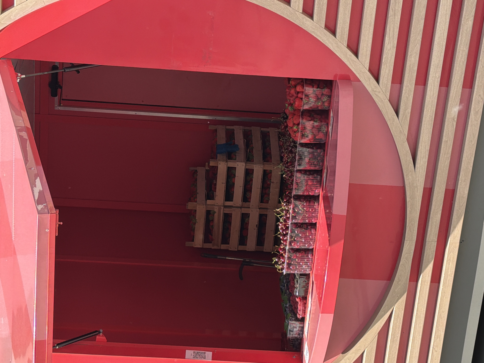 The image depicts the interior of a red, modern-looking vending machine or produce stand, likely situated in an outdoor or semi-outdoor public space in Moscow, Russia. The structure has a distinct rounded, elliptical opening, framed by a light-colored wooden archway that transitions into a background wall of alternating vertical wooden and red/pink striped panels.
Inside the vibrant red compartment, an abundant display of fresh berries is neatly organized. On the right side, multiple clear plastic containers filled with bright red strawberries are stacked high, some appearing to be in wooden crates, others directly stacked. Below the strawberries, similar containers hold dark red cherries. Further down and to the left, more wooden crates are visible, some also containing berries, including what appears to be blueberries. A long, dark handling tool or mechanism extends upwards from the bottom right of the interior, possibly for retrieving items. Gas struts or similar hydraulic arms are visible near the top and bottom left, suggesting a hinged opening mechanism for the structure.
No people are present in the image, but the arrangement of the produce implies an activity of selling or dispensing fresh fruit. The lighting is bright and even, indicating daytime. A small QR code is visible on a white label on the lower left interior wall of the structure. The overall aesthetic combines bold red colors with natural wood elements, creating an inviting and contemporary design for a fresh produce dispenser.