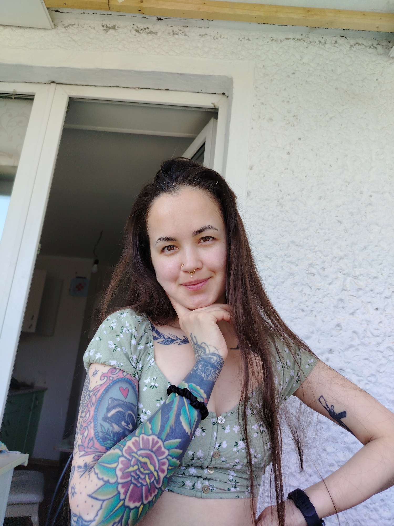 A young woman with long, dark hair is posing for a selfie. She is wearing a short-sleeved, floral-patterned green top. She has numerous tattoos on both arms, including a portrait of a raccoon. She is touching her chin with her right hand, and her left hand is resting on her hip. She is wearing a black watch on her left wrist and a black scrunchie on her right. The background includes a white door frame, a textured white wall, and a glimpse of an interior room. The location is likely in Noviy, Russia, based on the prompt.