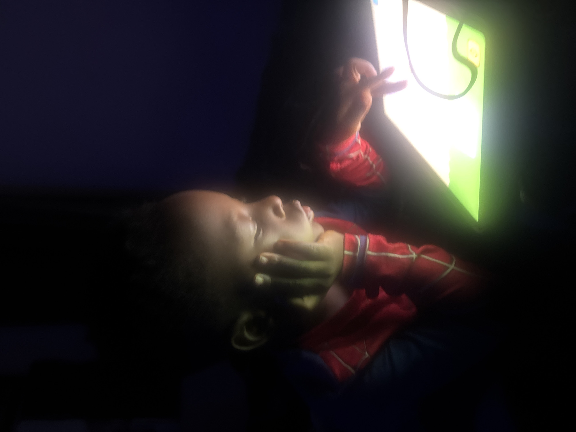 A young child, likely of African descent and wearing a red and blue suit, is seated in a dark room, illuminated by the bright screen of a tablet computer. The child's left hand is resting on their cheek, and their eyes are focused on the tablet. The child appears to be drawing or interacting with an application on the tablet, indicated by the finger touching the screen. The background is dark, suggesting the image was taken at night or in a dimly lit environment. The tablet's screen displays bright, colorful graphics, with a green area and a curved line visible. The scene takes place indoors, likely within a home in Gura Top, Nigeria.