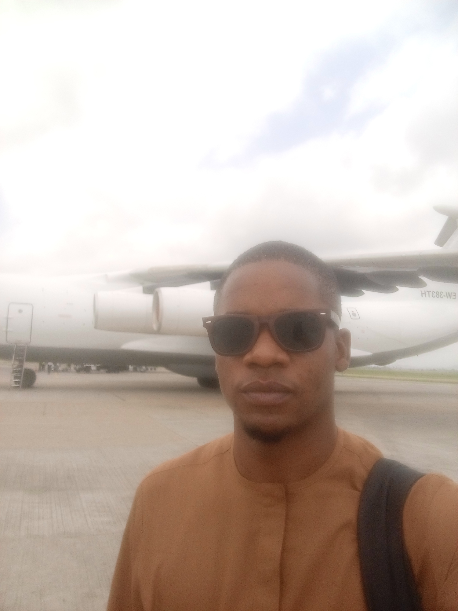 A man wearing sunglasses and a brown shirt stands in front of an airplane on a tarmac. The plane is white with the registration number HT-883-W3 visible on the tail. The environment appears hazy, suggesting a humid or overcast day. The location is Gwaska, Nigeria.