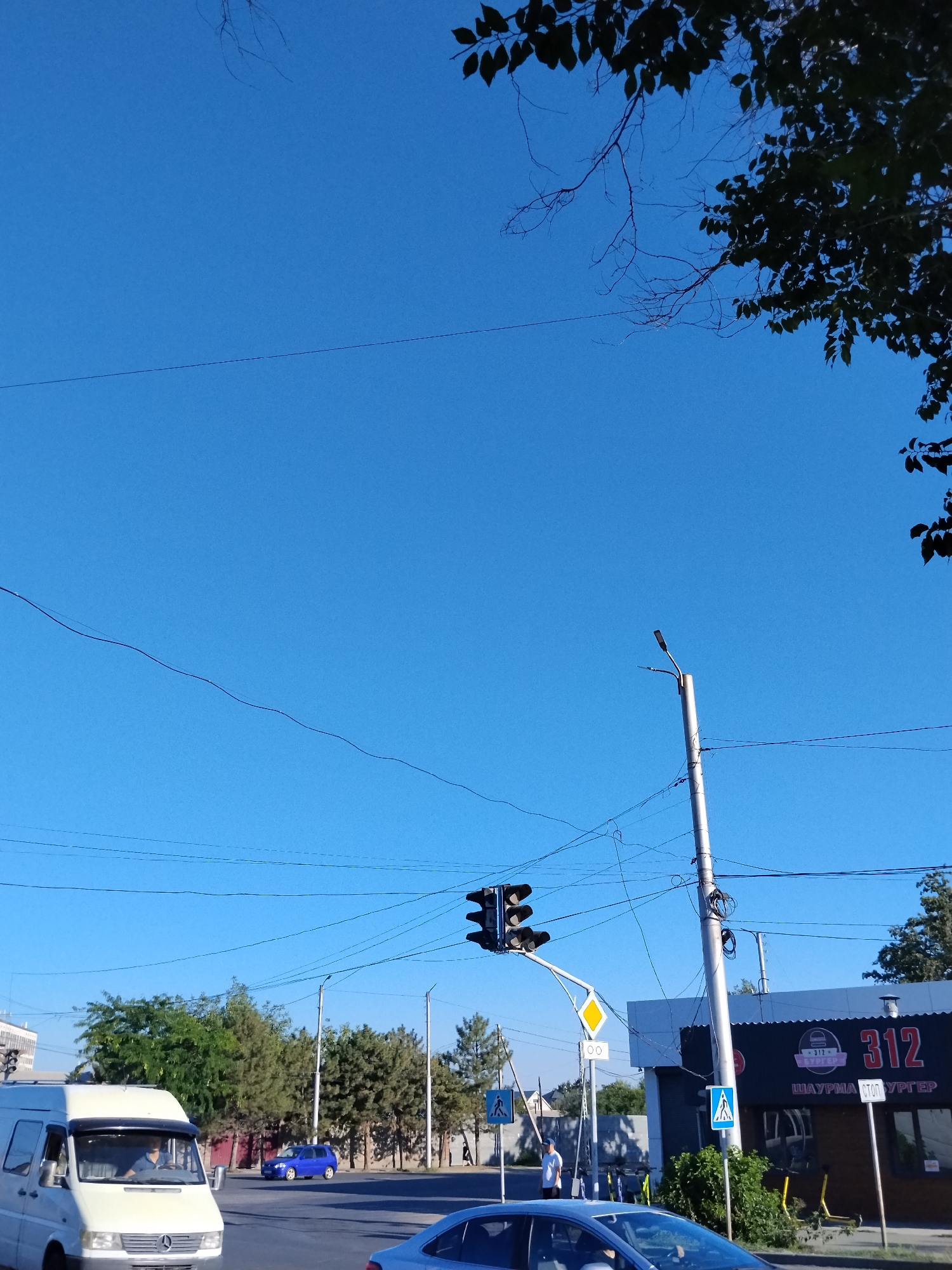 The image depicts a street scene in Bishkek, Kyrgyzstan, under a clear blue sky. In the foreground, a white van is partially visible on the left, with its driver behind the wheel. To the right, a silver car is in motion. Further ahead, a blue car is also on the road. Traffic lights are visible at an intersection, with one set showing a red light. Pedestrian crossing signs are posted on poles. A commercial building with the number "312" and the word "БУРГЕР" (burger) prominently displayed on its facade is situated on the right side of the street. Several trees line the background. Electrical wires and a street lamp are also present.