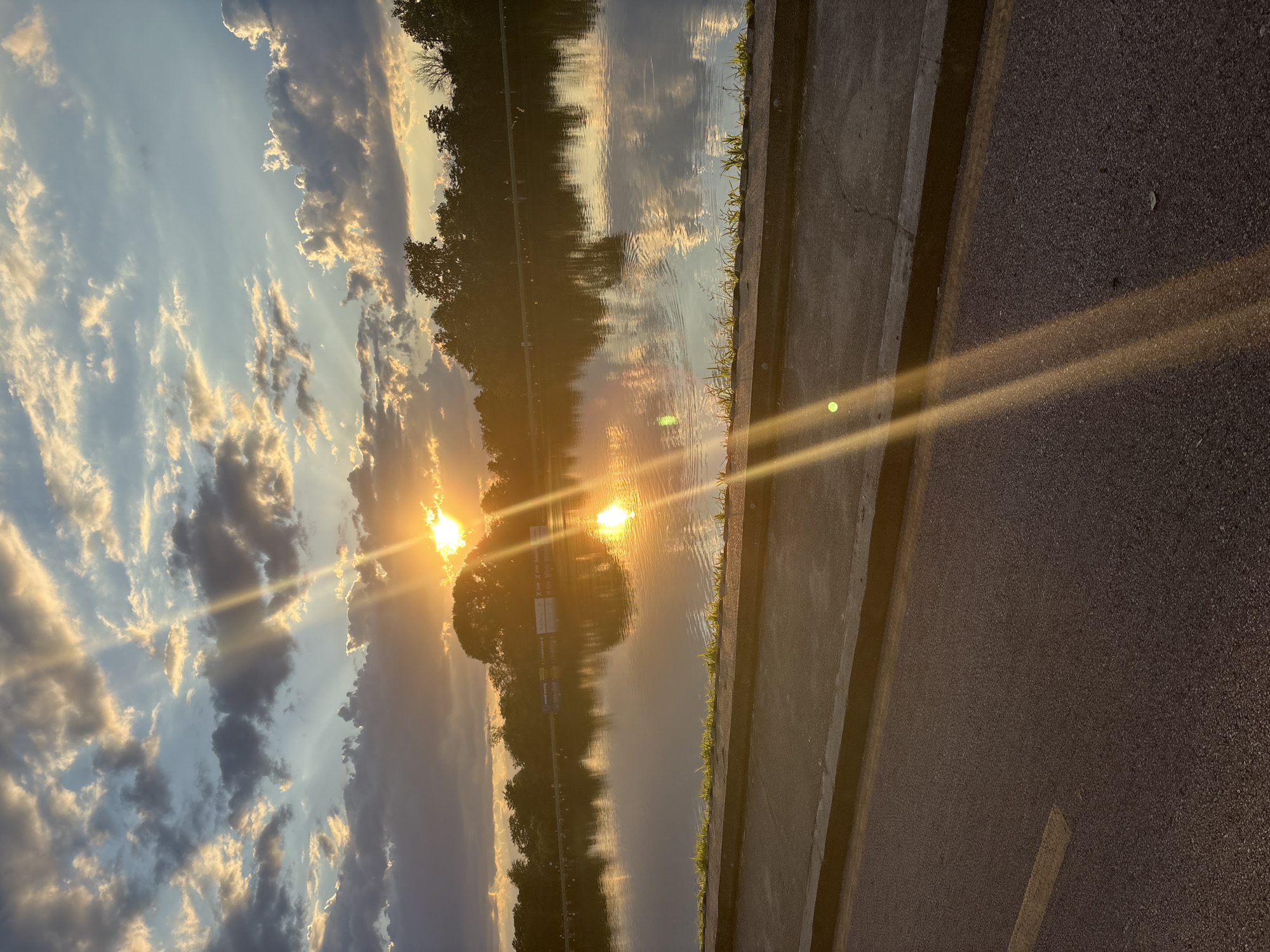 The image depicts a sunset over a body of water, likely a lake or river, with a road and embankment in the foreground. The sky is filled with dramatic clouds, illuminated by the setting sun, which casts golden rays across the scene. The water reflects the sky and trees lining the opposite bank. There is a paved road with a yellow line, a concrete curb, and some grass growing on the embankment. The overall environment suggests a park or recreational area. While the image is detailed, it does not contain specific visual cues that would allow for definitive identification of its location within the city of Plovdiv, Bulgaria.