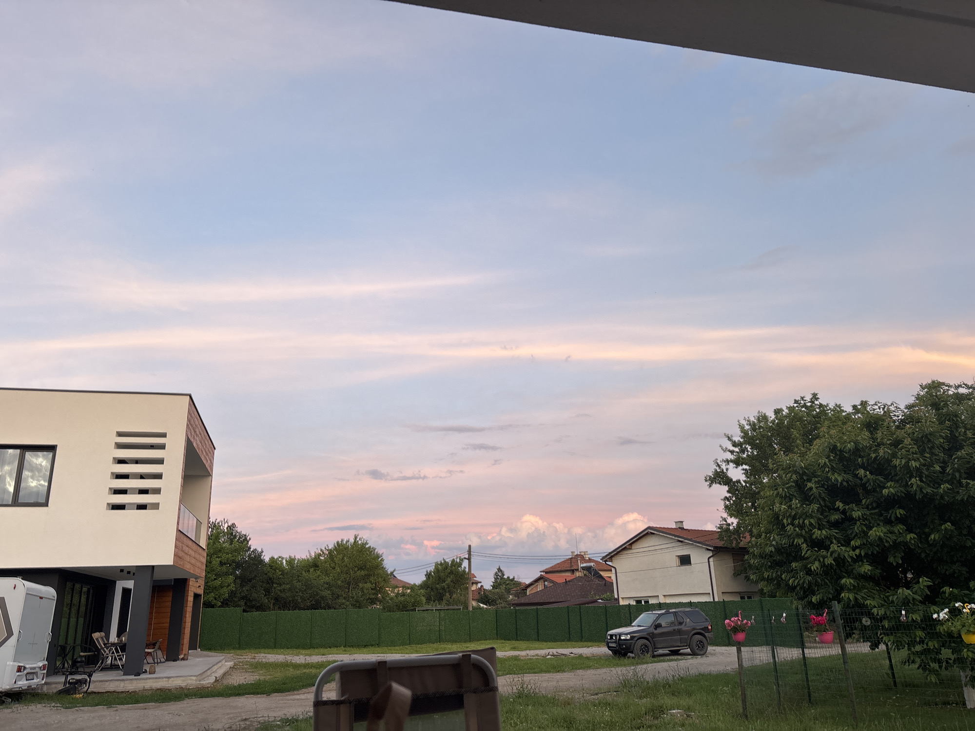 The image captures a residential scene in Mramor, Bulgaria, during what appears to be sunset. The sky is painted with soft hues of pink and blue, with wispy clouds stretched across the horizon. In the foreground, a modern, multi-story building with white and wood-paneled facade is visible on the left. Adjacent to it, a white recreational vehicle is parked. A dark-colored SUV is positioned on a gravel path in front of a lush green fence that encloses the property. Beyond the fence, several houses with red-tiled roofs are nestled amongst trees. A large, dense tree dominates the right side of the frame, its branches reaching towards the sky. There are no people visible in the image. The overall impression is one of a quiet, suburban evening.
