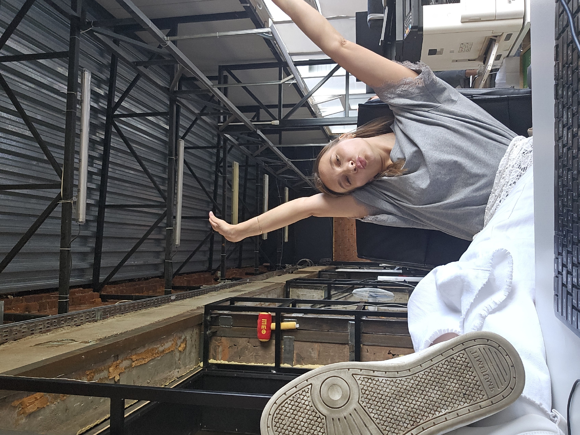 A young woman is captured posing playfully in an industrial or workshop-like indoor environment in Chaikovsky, Russia. She is horizontally oriented in the frame, wearing a grey t-shirt with lace details on the sleeves and white pants or skirt with lace trim. Her arms are outstretched, and she has a pouting or kissy expression.
The setting features extensive black metal scaffolding and framework, with corrugated metal walls forming the background. A deep channel or pit runs through the foreground and midground, revealing exposed brickwork and orange foam insulation along its sides. Long, fluorescent-like light tubes are attached to the metal structures.
On the right side of the image, a computer keyboard is partially visible, along with a white printer or copier machine in the background. A small white fan is also seen on a surface. A red and yellow object, possibly a toy hammer or prop, rests near the channel with the Cyrillic text "ТШФ" clearly visible. The sole of the woman's light-colored sneaker is prominent in the bottom right, showing a textured pattern and partially visible text, possibly "TIMOIMU", indicating a brand name. The lighting suggests it is daytime.