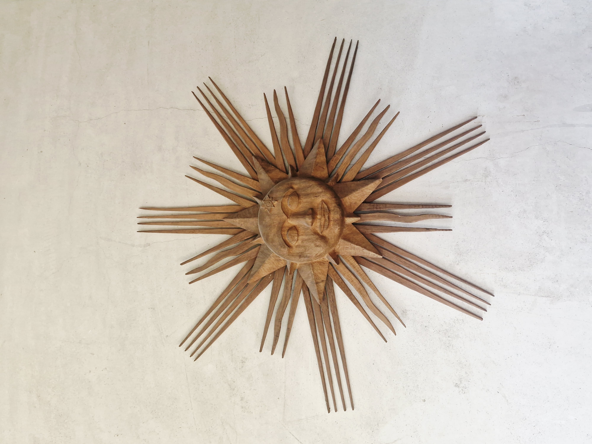 A wooden sun decoration is centered against a neutral, off-white surface. The sun is crafted from wood and features a central circular face with carved facial features. From the central face, wooden rays extend outwards, some thicker and more triangular near the center, and some thinner and more pointed, resembling elongated spikes. The sun decoration is textured and appears to be carved by hand. The surface underneath is plain and uniformly colored, with subtle variations in tone and possible markings. The image captures a static, overhead view of the sun decoration against the simple background. There is no indication of this photo being taken in Hrabrino, Bulgaria, or any other specific location.