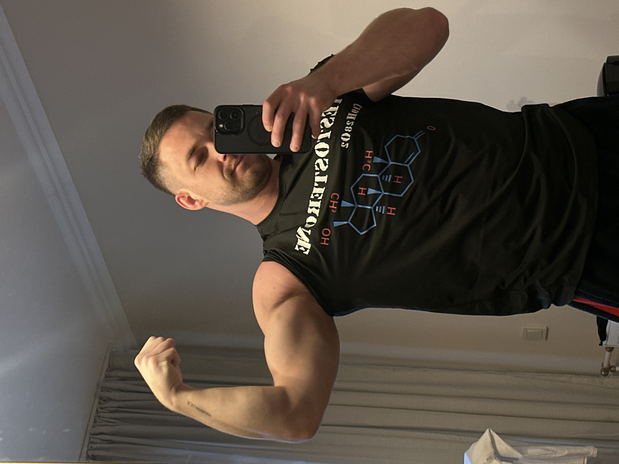 A man with short brown hair and a light beard is standing indoors and taking a selfie in a mirror. He is wearing a black t-shirt with a chemical formula of testosterone on it. He is flexing his left bicep. The location is not specified in the city of Perm, Russia, and cannot be determined from the image.