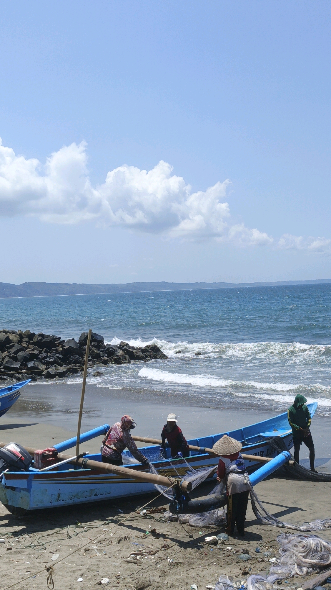 The image depicts a coastal scene at Desa Pagandaran, Indonesia, on a bright, sunny day with scattered clouds. In the foreground, a sandy beach is littered with fishing nets, debris, and two blue fishing boats. One boat is pulled onto the sand, while another is partially visible on the left.
Within the blue boat on the sand, three individuals are present. One person, wearing a patterned hoodie and a head covering, is seated and appears to be tending to fishing nets. Another person, wearing a white cap and a dark shirt, is also seated in the boat. A third person, wearing a conical hat and a light-colored shirt with dark pants, stands behind the boat, interacting with the fishing nets. To the right of this boat, another person wearing a green hoodie stands on the sand.
The ocean is a vibrant blue with moderate waves breaking on the shore. A rocky breakwater is visible on the left side of the image, separating the beach from a stretch of calm water. In the background, a hazy, mountainous coastline stretches across the horizon under a pale blue sky. The overall scene suggests a fishing community preparing or finishing their work at the seaside.