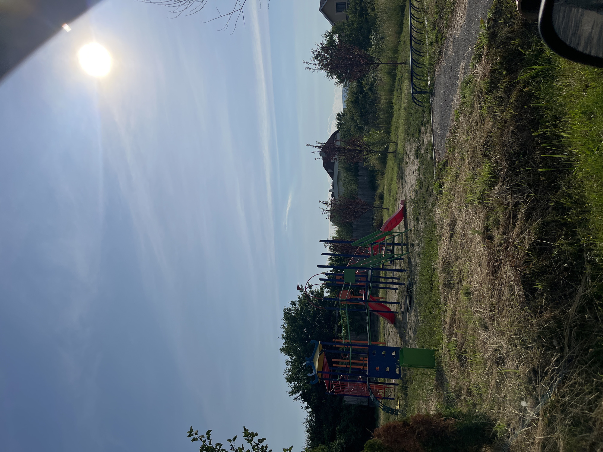 A bright, sunny day is depicted in this outdoor scene, likely a public park or playground, located in the city of Onokivtsi, Ukraine. The sky is a clear, pale blue with a few wispy white clouds. The sun is prominently visible in the upper right corner, appearing as a bright white orb, casting a glare and lens flare.
In the mid-ground, a colorful playground structure stands on a sandy surface. The structure is made of metal and plastic, with red, blue, green, and orange elements. It features slides, a climbing frame, and platforms. A green trash bin is situated to the left of the playground. Beyond the playground, a wooden fence runs horizontally across the scene. Several small trees with red-tinged leaves are planted in a grassy area. In the background, houses with red-tiled roofs are visible, nestled amongst trees.
The foreground and mid-ground are covered in dry, brown grass interspersed with patches of greener grass. A gravel pathway curves through the scene, leading towards a bicycle rack. The overall impression is one of a quiet, functional outdoor space on a pleasant day. There are no people visible in the image.