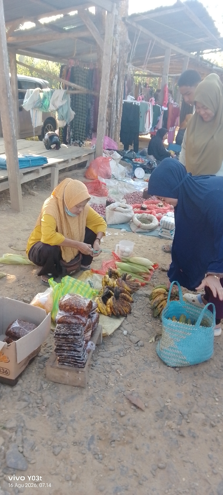This image depicts an outdoor market scene in Palosilae, Indonesia. The market is set up under a rustic, open-air shelter constructed with wooden poles and a corrugated metal roof. In the foreground, a woman wearing a yellow hijab and yellow shirt is crouched down, appearing to be handling some items, possibly money or goods, in her hands. In front of her, a collection of packaged food items, stacked neatly, are placed on a wooden plank. To her right, a bunch of bananas and several ears of corn are displayed on a yellow tarp. Further to the right, a blue woven basket contains more bananas. In the background, a variety of produce, including onions and tomatoes, are spread out on a white tarp. Several people, including a woman in a dark hijab and another in a light brown hijab, are visible interacting around the market stalls. Clothing items are also displayed on racks under the shelter. The ground appears to be sandy or dirt. A car is partially visible in the left background. The overall scene suggests a bustling local market environment.